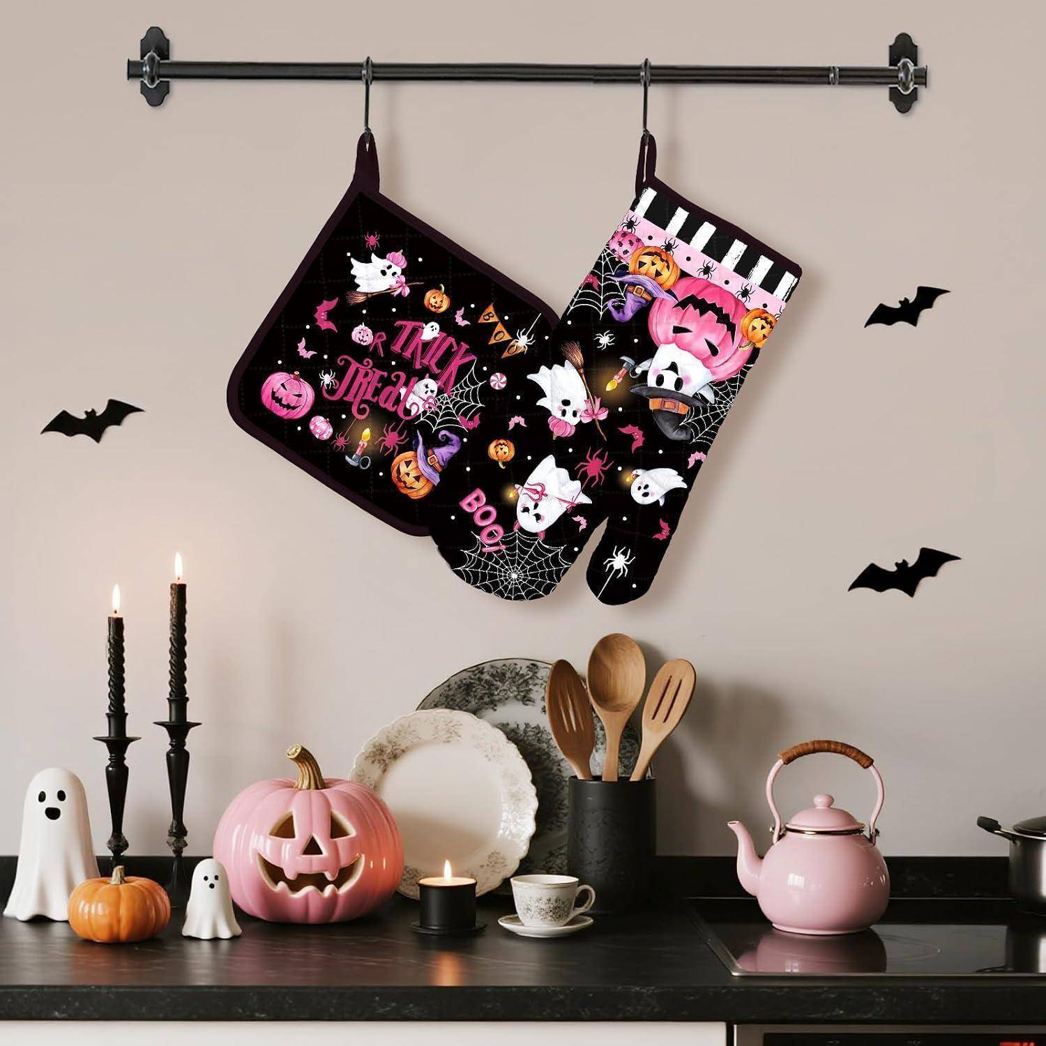 Halloween-Küchendisplay mit zwei hängenden Ofenhandschuhen, Fledermaus-Wandaufklebern, brennenden Kerzen, Kürbissen, Geistfiguren und einer rosa Teekanne auf der Arbeitsplatte.