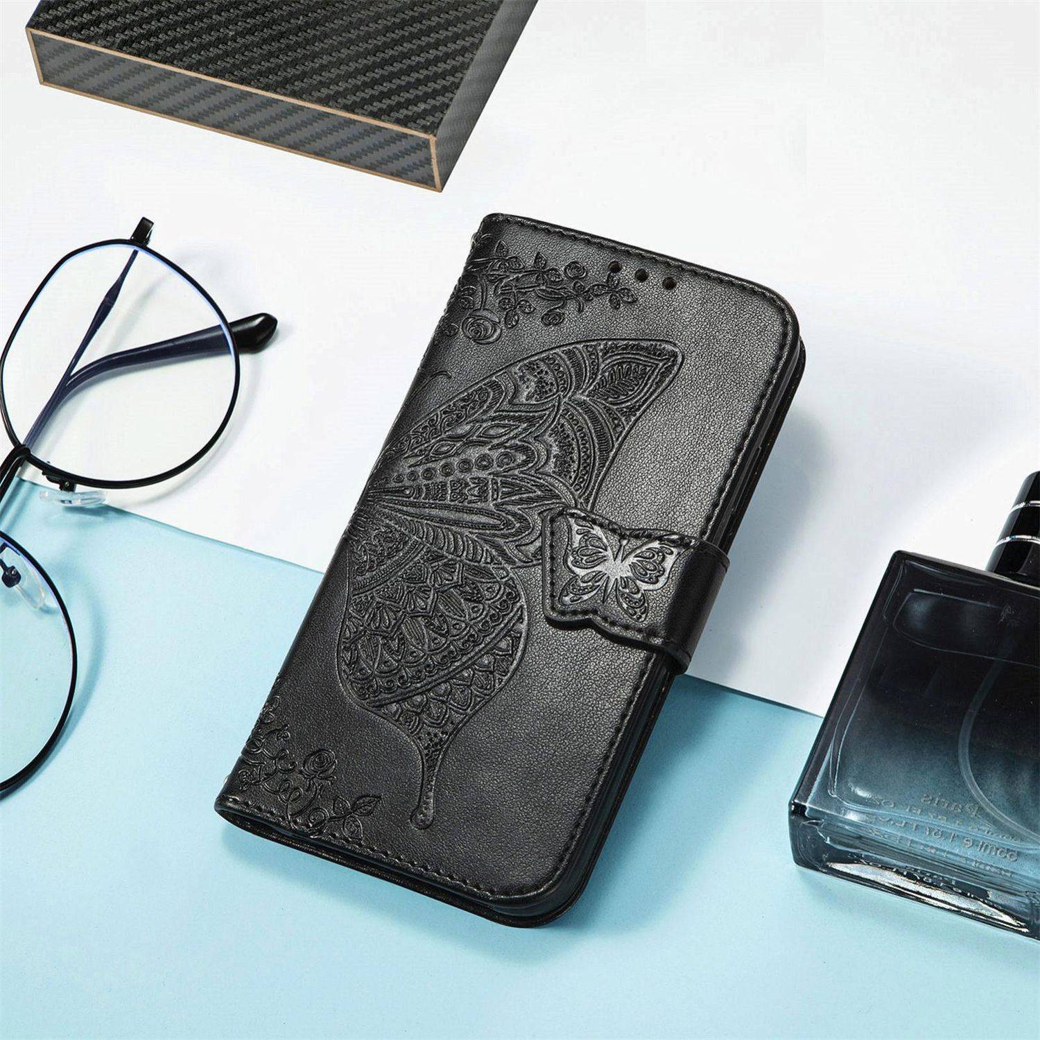 Schwarze geprägte Schmetterlings-Handytasche auf einer hellblauen und weißen Oberfläche, neben einer Brille und einer Parfümflasche.
