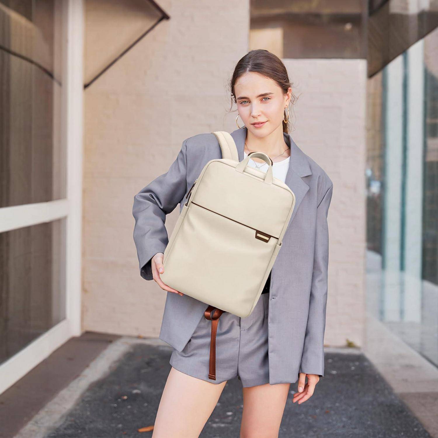 Frau in einem grauen Anzug, die einen cremefarbenen Rucksack im Freien in einer urbanen Gehweg hält und die Tasche modelliert.
