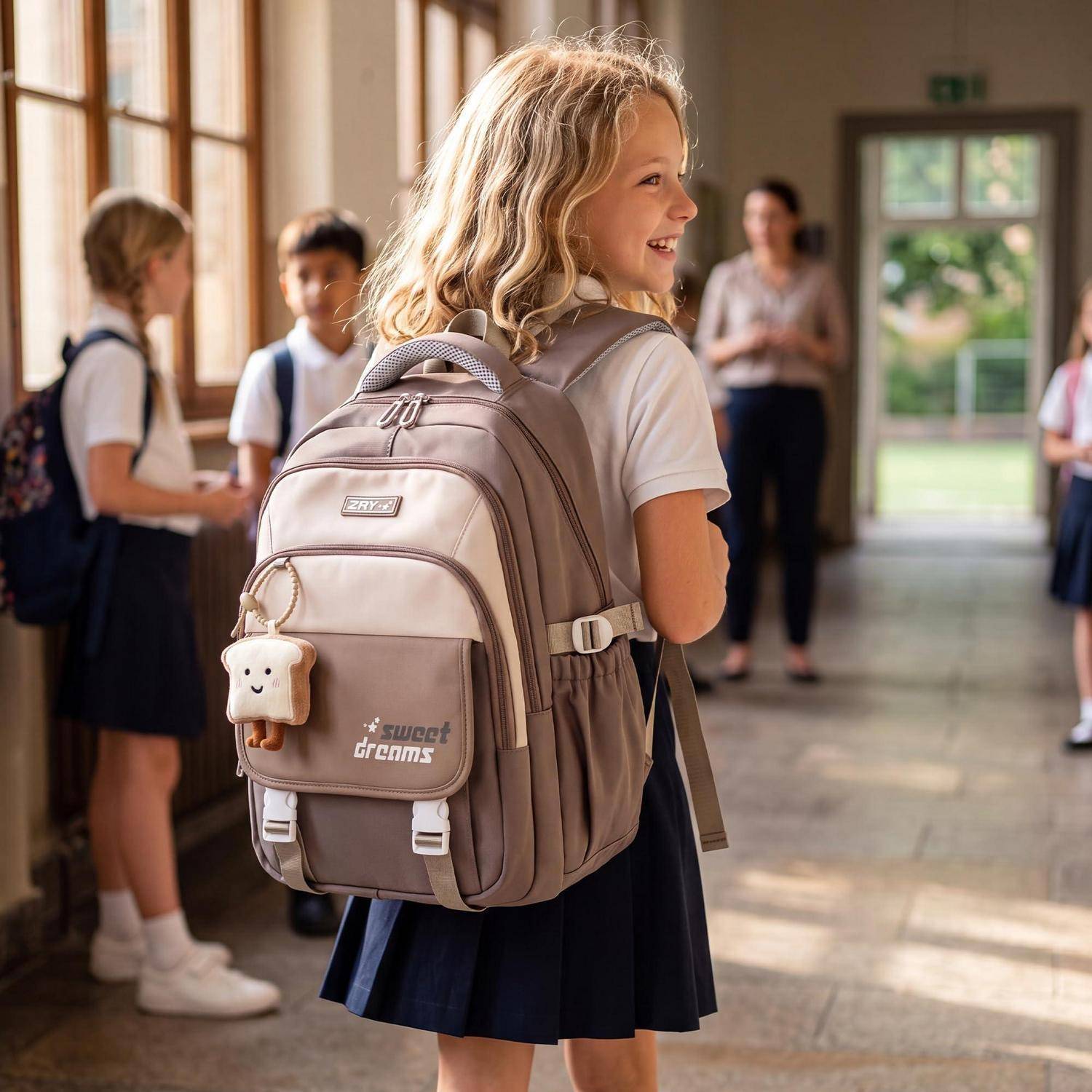 Lächelndes Schulmädchen mit einem großen Rucksack in einem Schulflur, mit anderen Kindern und einer Lehrerin im Hintergrund.