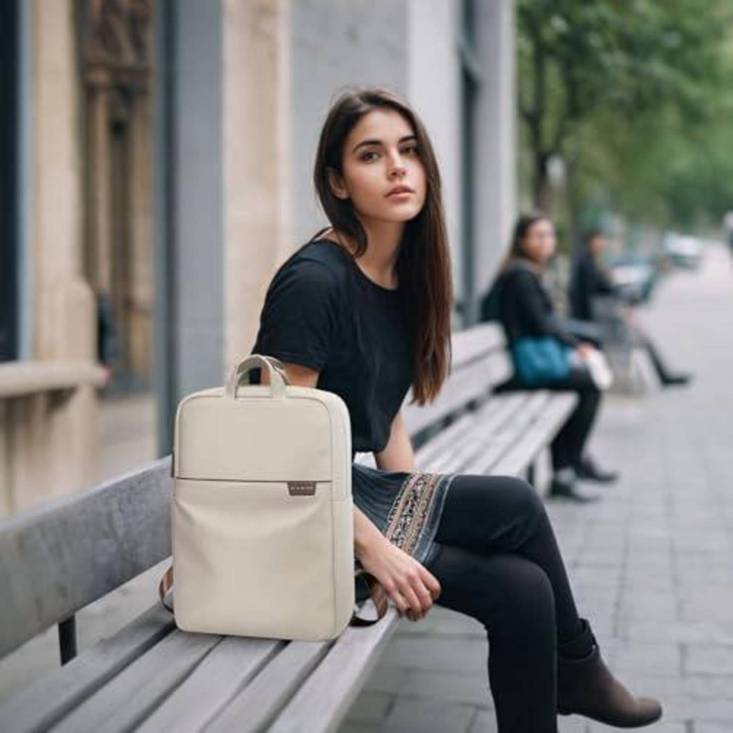 Junge Frau in Schwarz, die auf einer Bank sitzt, mit einem beigen Rucksack neben sich auf einem Gehweg der Stadt, schaut in die Kamera.