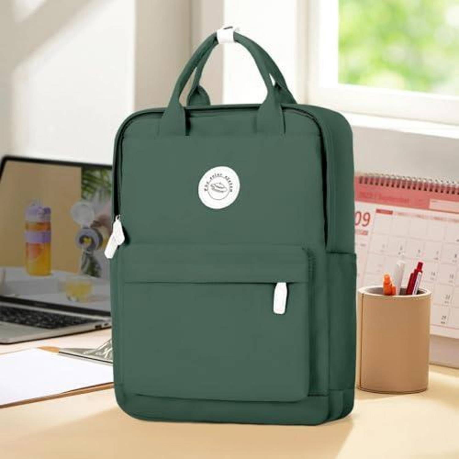 Grüner Rucksack mit Vordertasche und Griffen oben auf einem Schreibtisch neben einem Laptop, Kalender und Stifthalter.