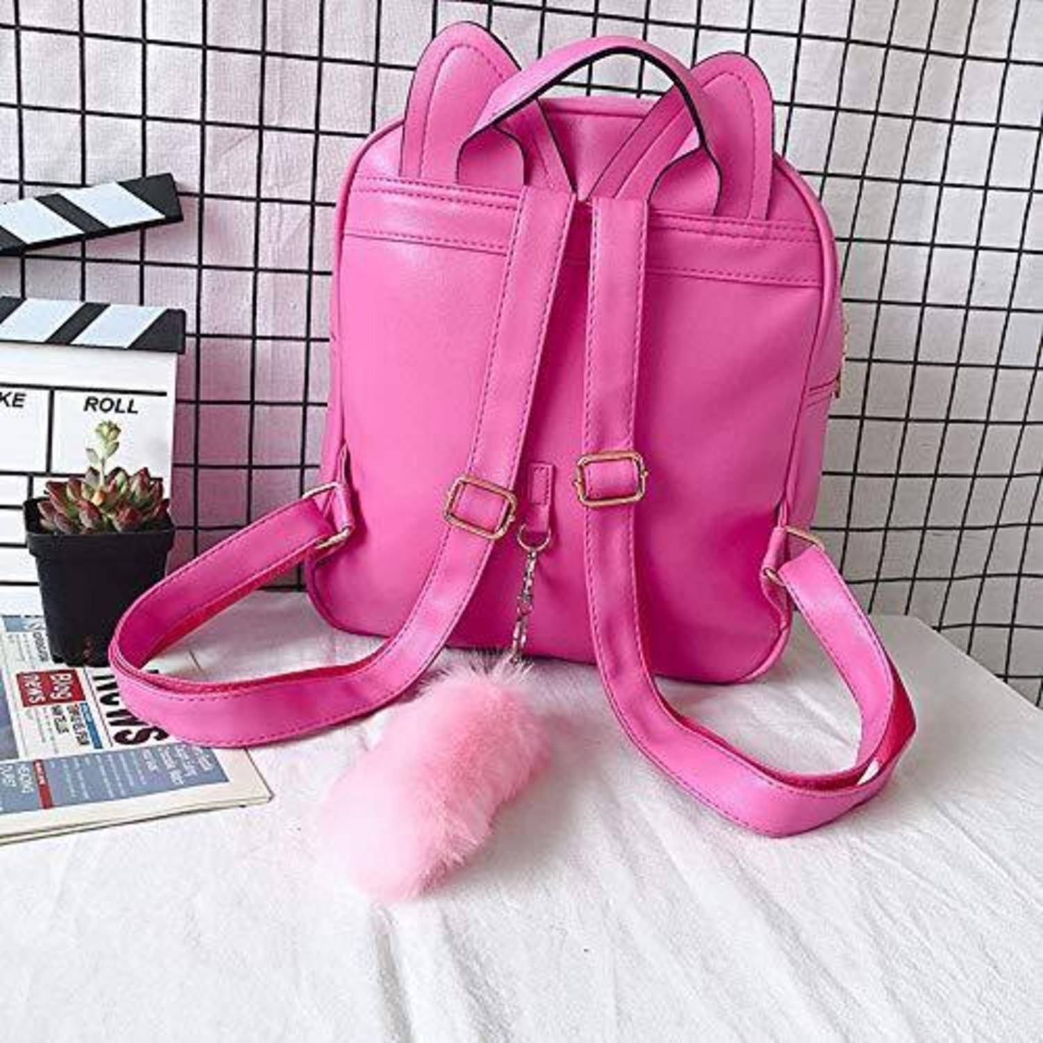 Leuchtend rosafarbener Rucksackbeutel mit vorderen Schnallen und flauschigem Pompom-Anhänger auf einer weißen Oberfläche neben einer kleinen Pflanze und einer Filmklappenbrett-Requisite.