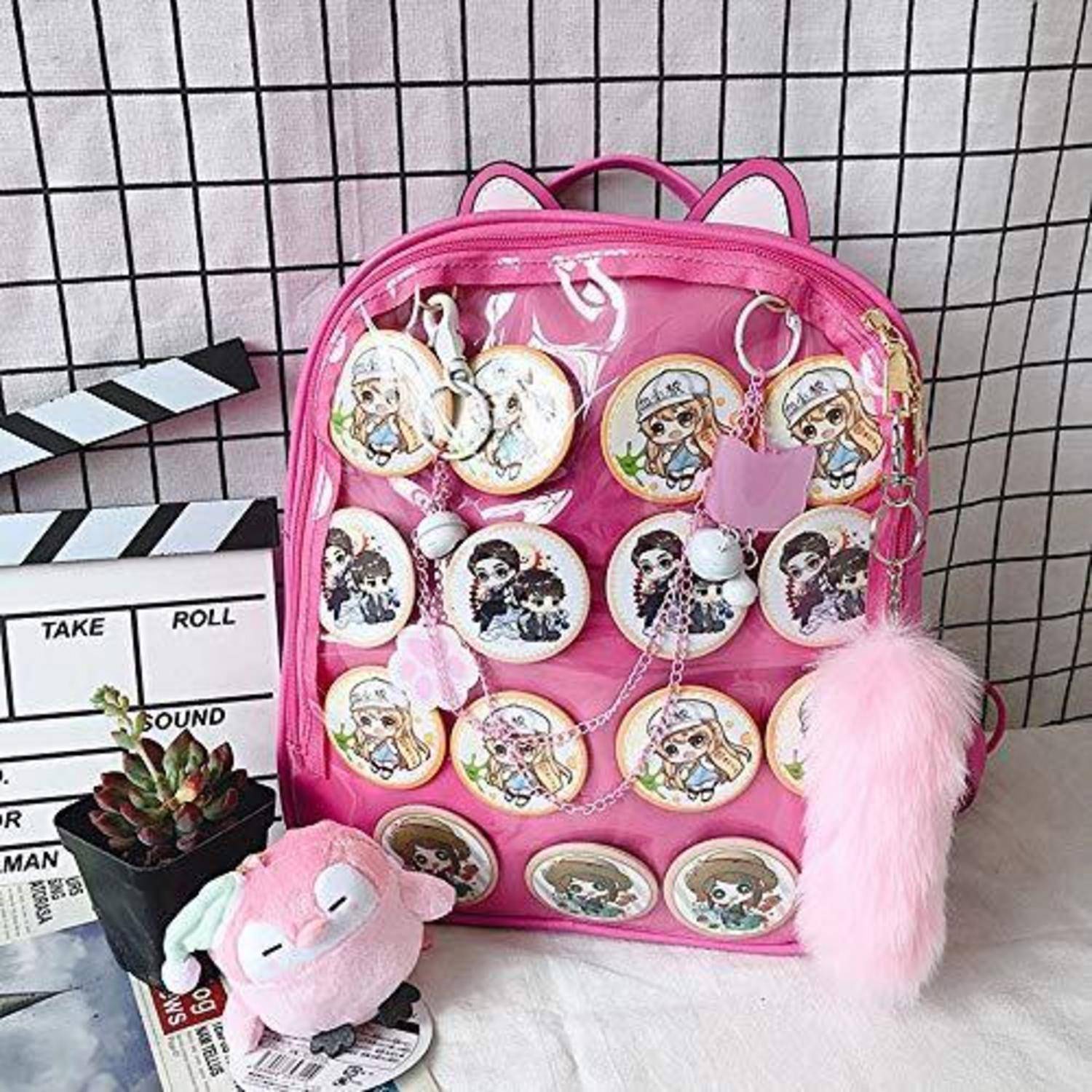 Rosarucksack mit Cartoon-Charakter-Pins, einem flauschigen Schlüsselanhänger und Bommel, neben einer Filmklappe und einer kleinen Topfpflanze ausgestellt.