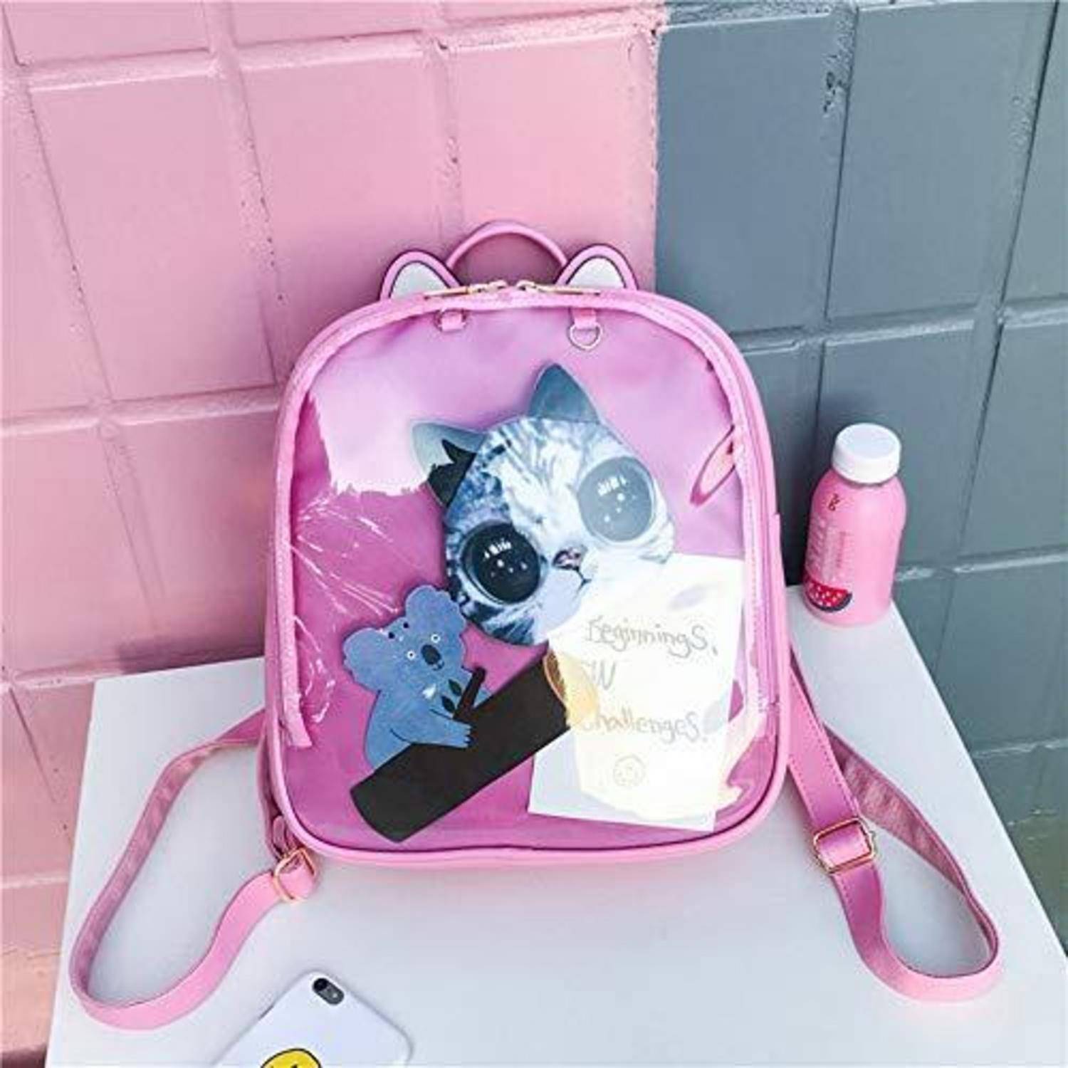 Rosa katzenmotivierter Rucksack mit einem Cartoon-Tiergesicht und blauer Bärengrafik auf einem weißen Tisch, neben einer rosa Flasche und einem Telefon.