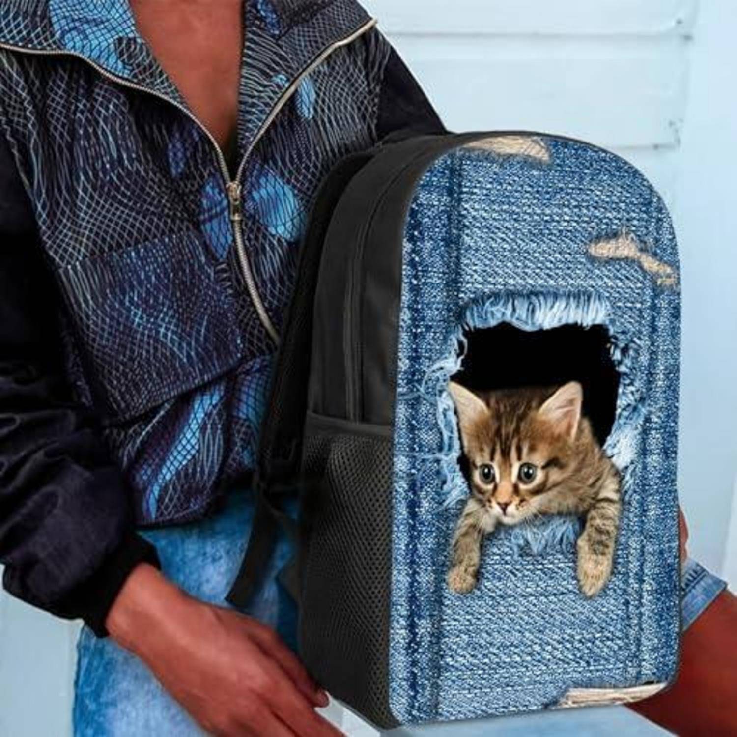 Person, der einen Rucksack-Transportbehälter im Denim-Stil hält, aus dessen vorderer Öffnung ein Kätzchen hervorguckt.