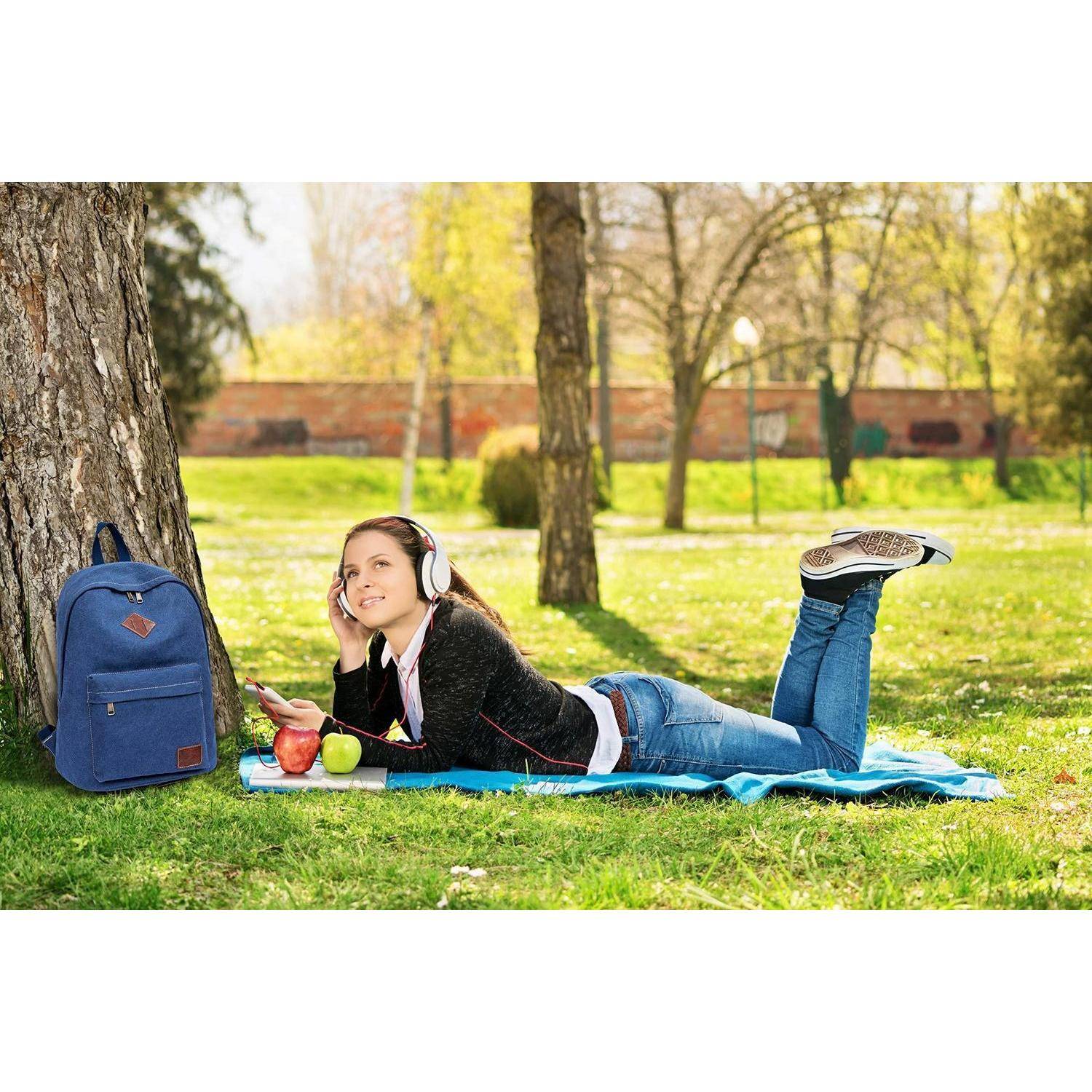 Junge Frau, die auf einer Decke im Park liegt, Musik mit Kopfhörern hört und ein Telefon neben einem Rucksack, Äpfeln und einem Buch benutzt.
