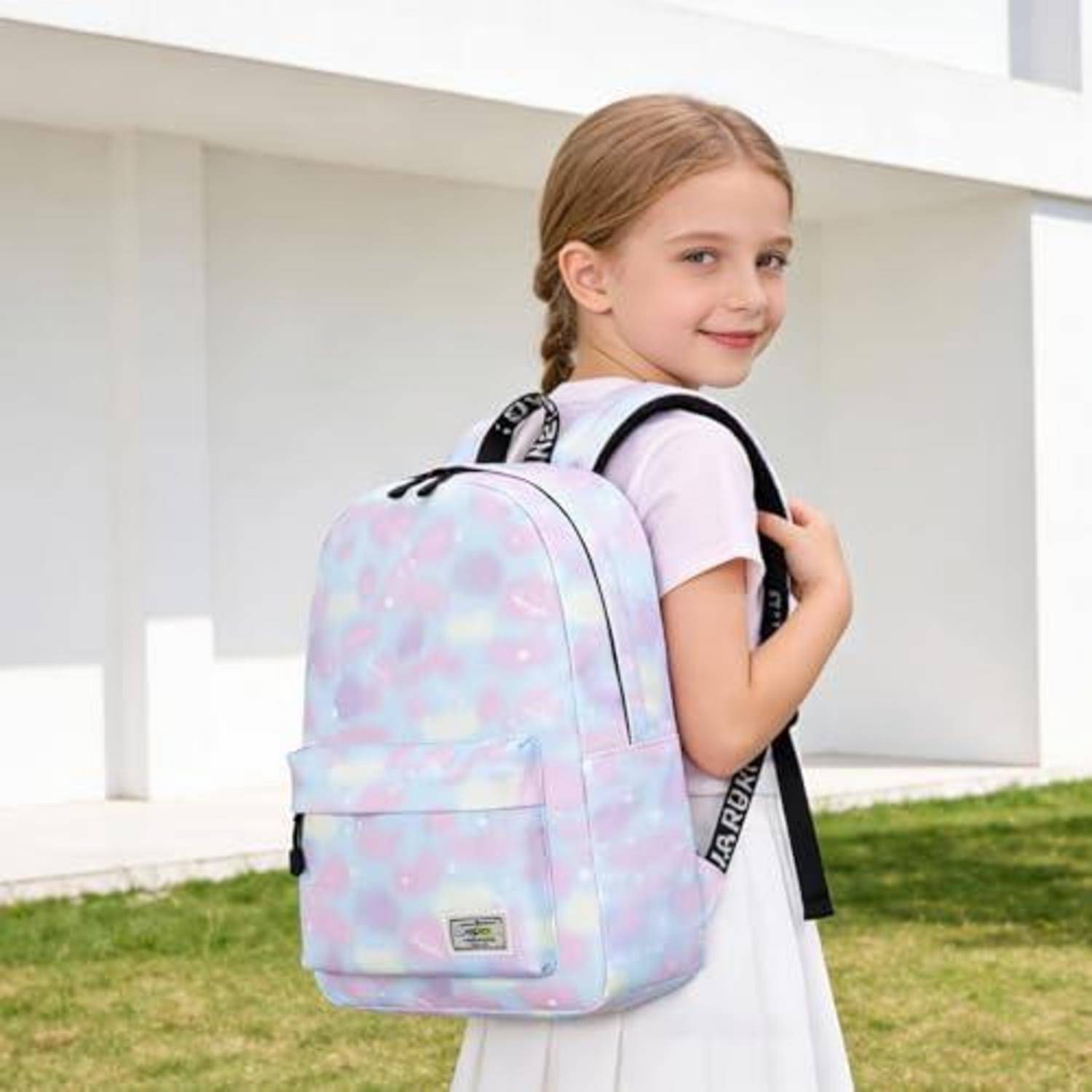 Junges Mädchen mit einem Pastellrucksack, das lächelt und draußen neben einem weißen Gebäude steht.