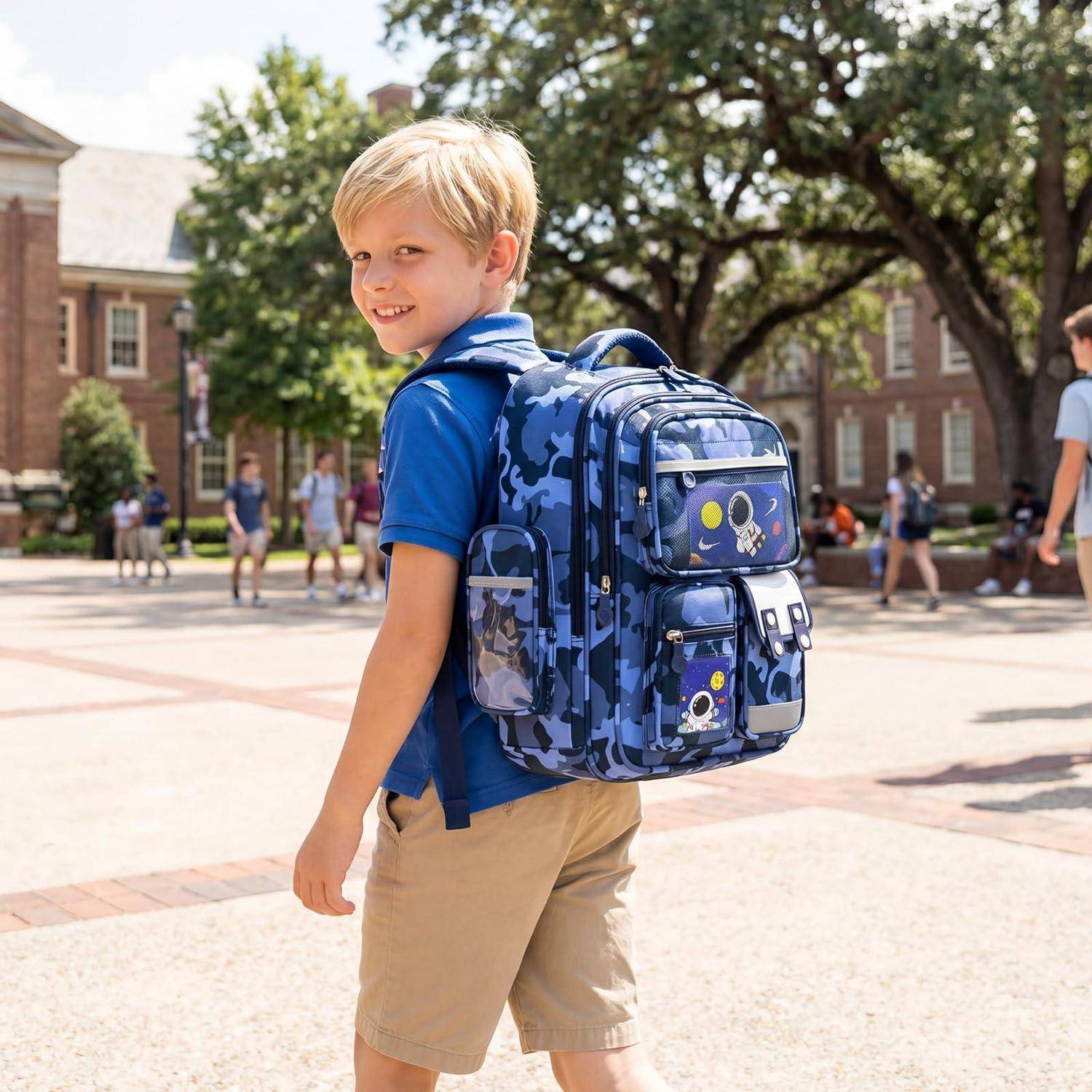 Junger Junge in blauem Hemd und khakifarbenen Shorts mit einem großen blauen Rucksack, der auf einem Schulgelände steht und über die Schulter in die Kamera schaut.