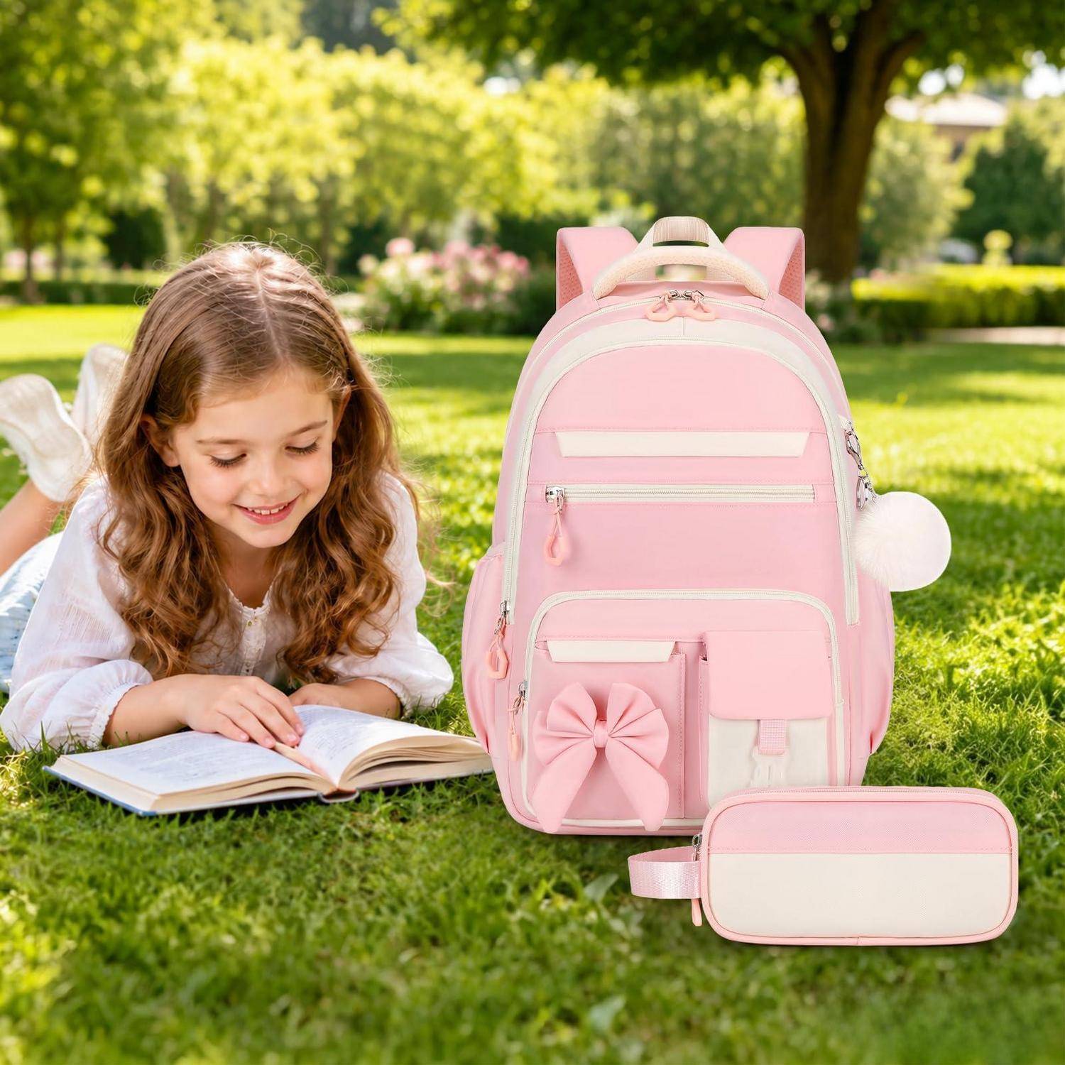 Junges Mädchen, das auf dem Gras neben einem rosa Schulrucksack und einer passenden Lunch-Box in einem sonnigen Park liest