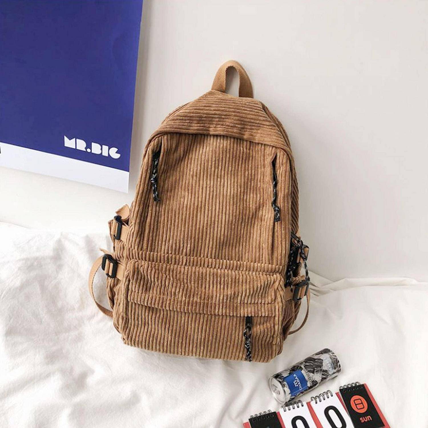 Brauner Kordsamtrucksack auf einem weißen Bett, mit einer kleinen Getränkedose und einem Notizbuch daneben.
