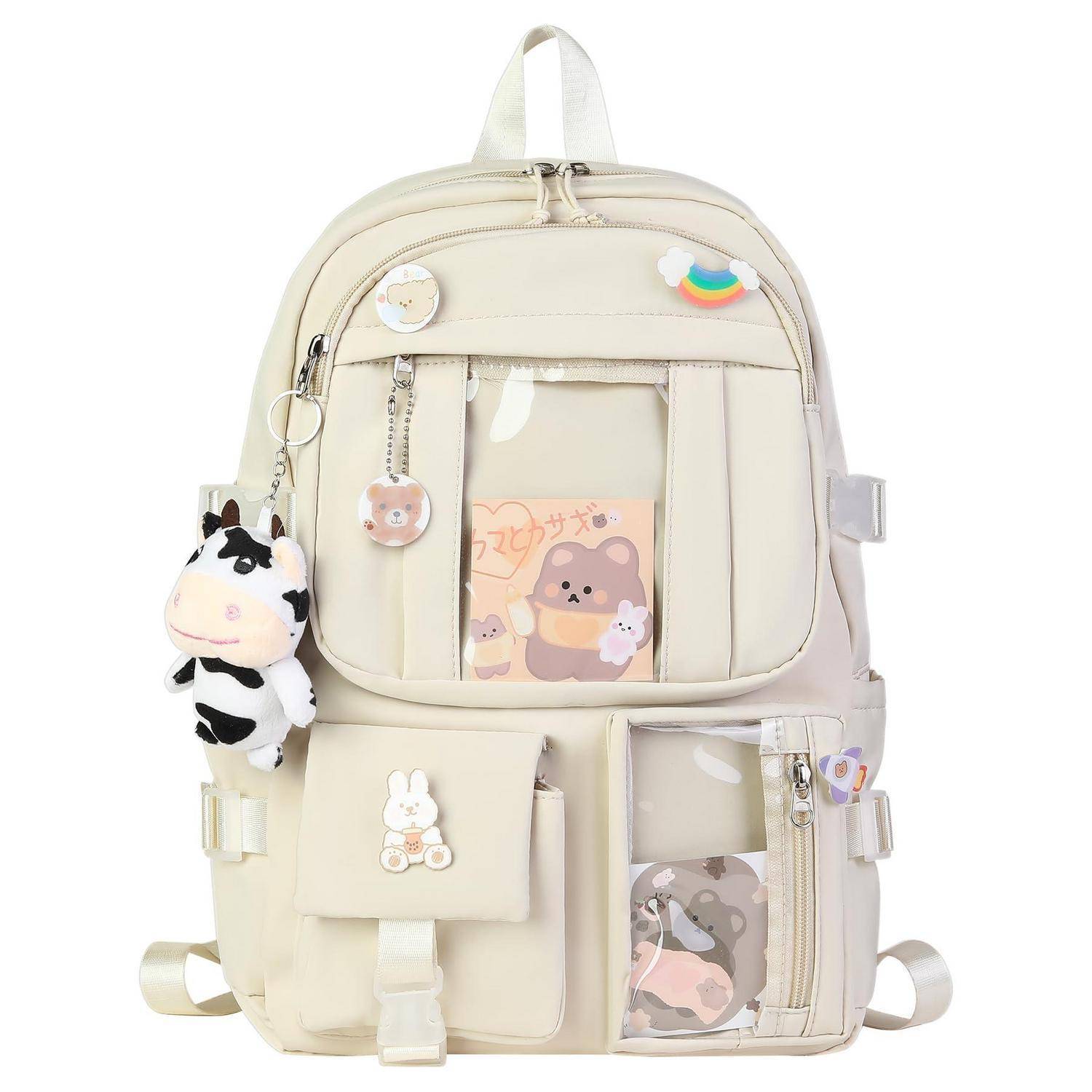 Cremefarbener Rucksack mit niedlichen Cartoon-Anhängern verziert, einer vorderen Tasche und kleinen Reißverschlussfächern auf weißem Hintergrund.