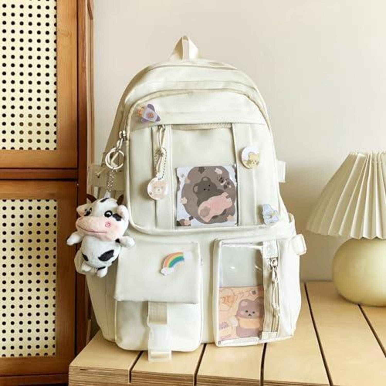 Cremefarbener Rucksack mit mehreren Vordertaschen und niedlichen Cartoon-Ansteckern, einschließlich eines Plüsch-Kuh-Charms und eines Regenbogen-Patches, auf einem Holztisch neben einer Lampe ausgestellt.