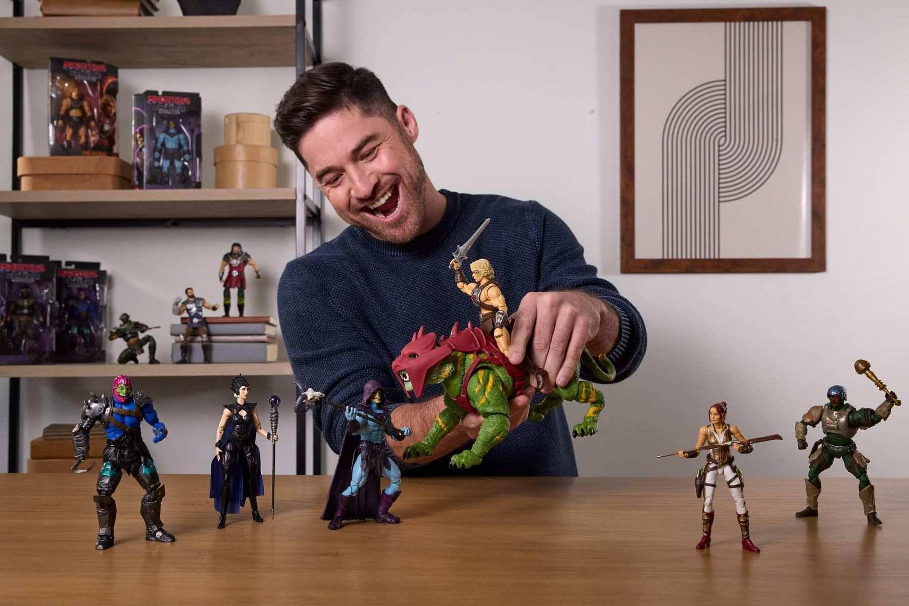 Ein lächelnder Mann spielt an einem Tisch mit Actionfiguren und arrangiert farbenfrohe Spielzeugfiguren in einem Wohnzimmer mit Regalen und Wandkunst.