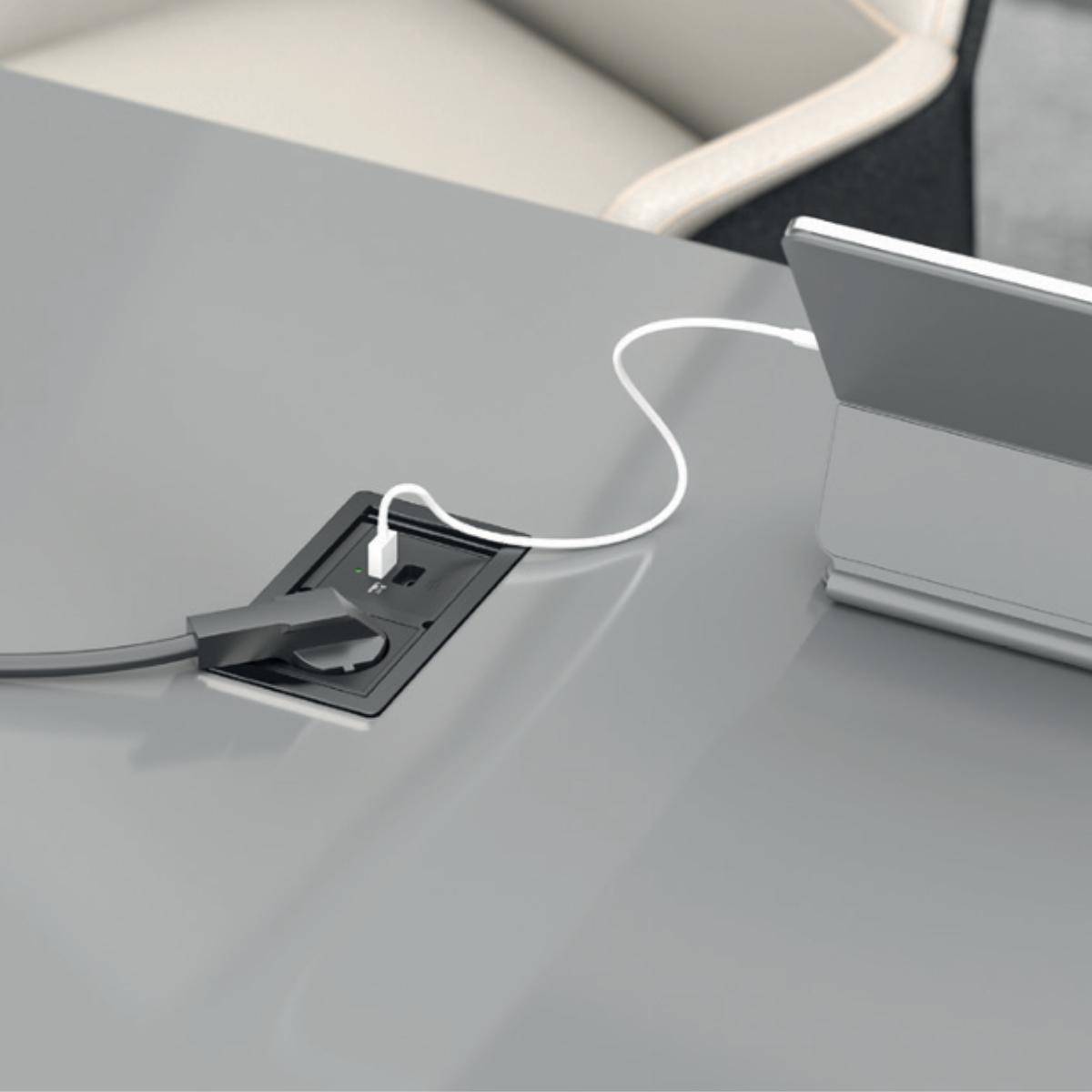 Tisch mit eingebautem Stromanschluss und Kabelmanagement, auf dem ein weißes USB-Kabel angeschlossen an ein Geräteladegerät zu sehen ist.