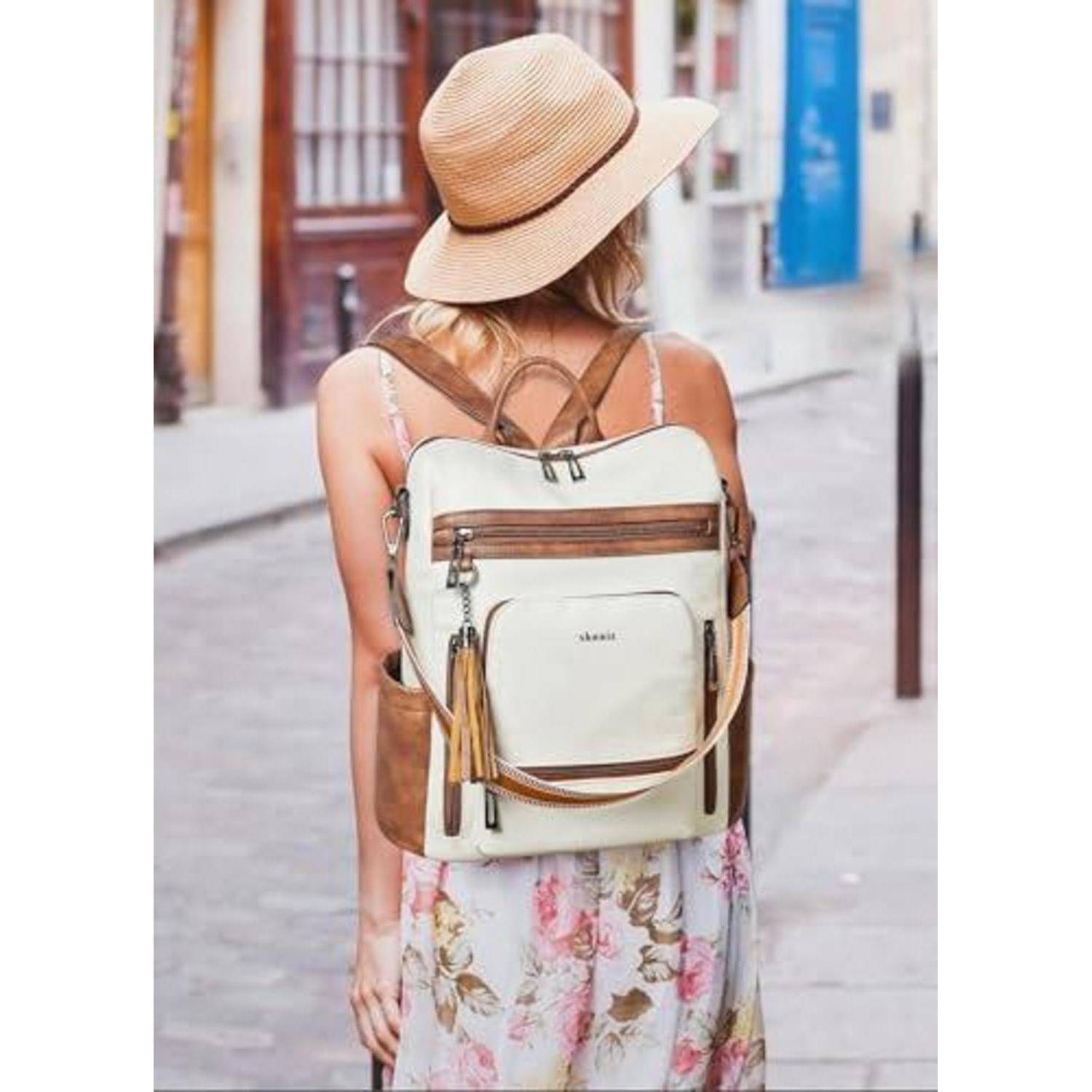 Eine von hinten zu sehende Frau, die einen Strohhut, ein Blumenkleid und einen cremefarbenen Rucksack trägt, während sie eine Straße hinuntergeht.