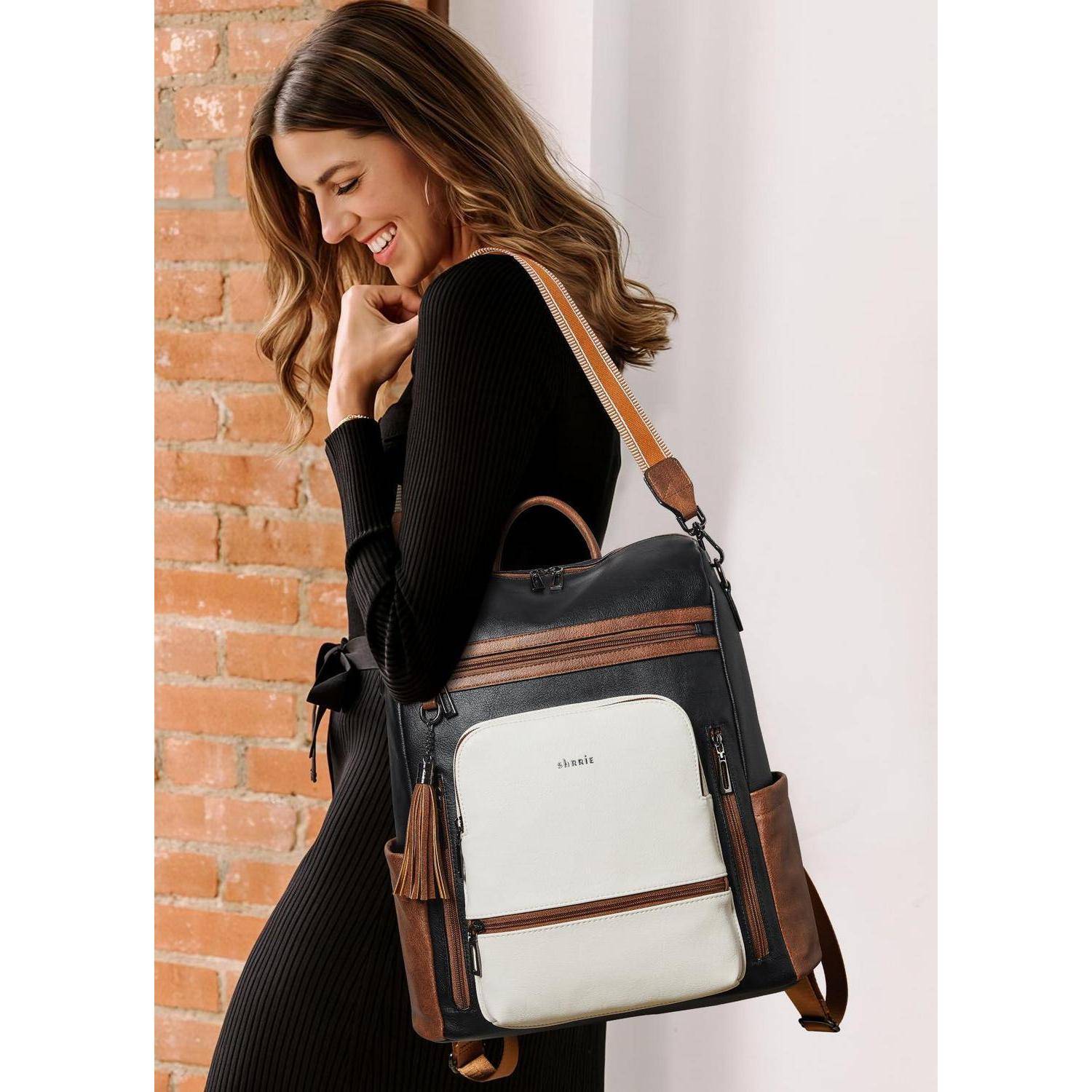 Frau in schwarzem Kleid, die einen schwarzbraun gemusterten Rucksack mit weißer Vordertasche trägt, vor einer Backsteinmauer und weißem Vorhang fotografiert.