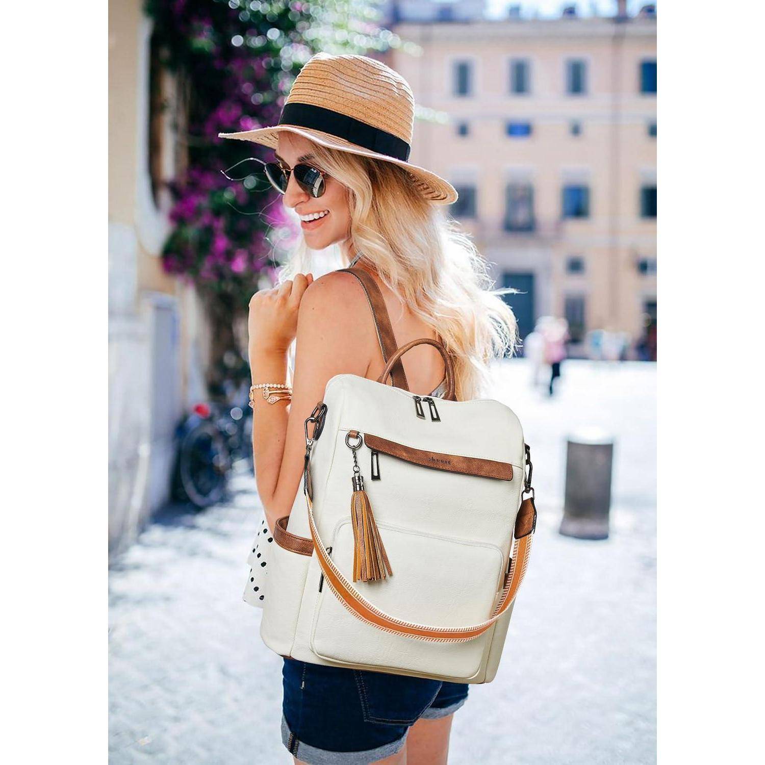 Lächelnde Frau in einem Strohhut mit Sonnenbrille trägt eine weiße Rucksack-förmige Handtasche, während sie an einem sonnigen Stadtstraße spazieren geht.