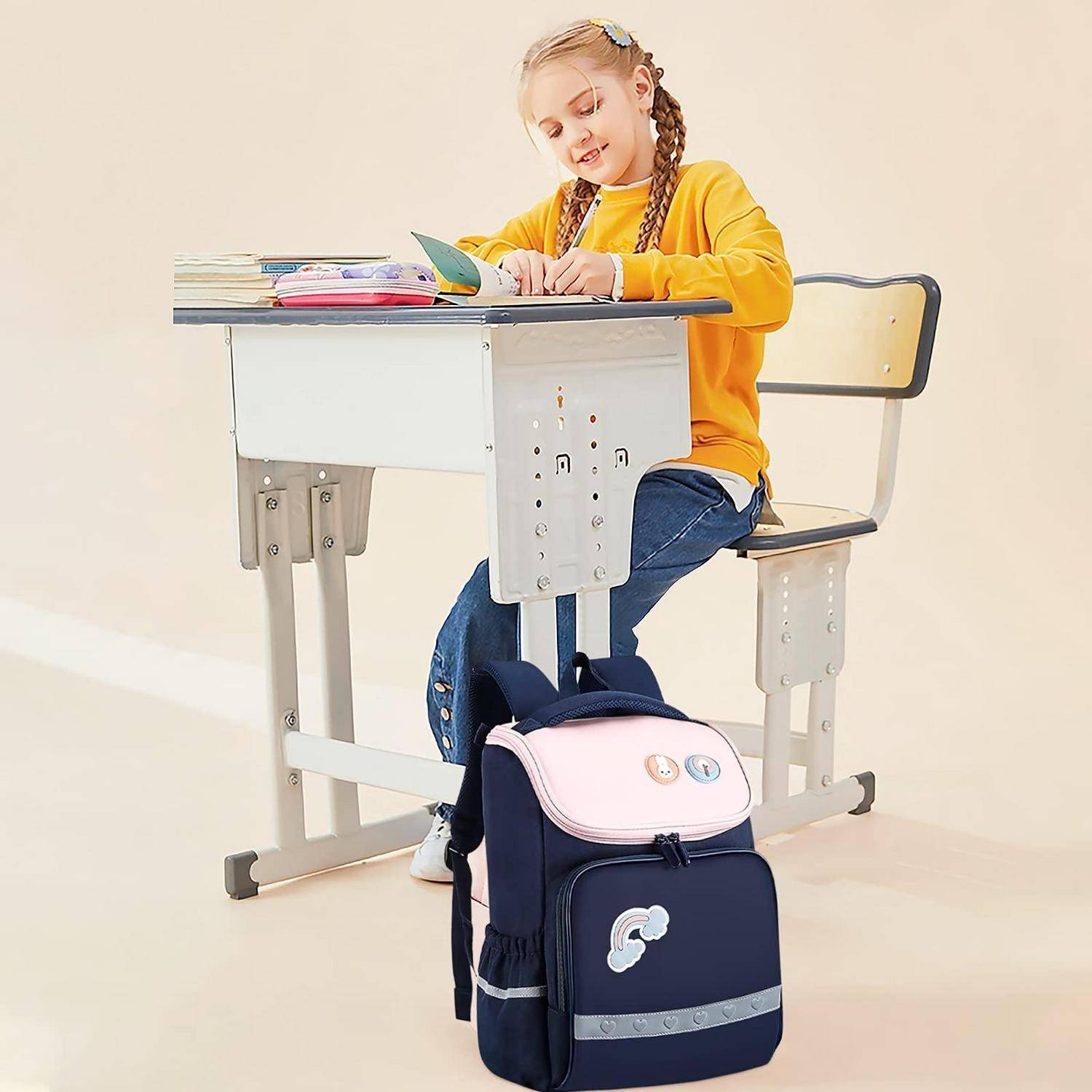 Junges Mädchen mit Zöpfen sitzt an einem Schultisch und schreibt in ein Notizbuch, während ein Rucksack über der Stuhllehne hängt.