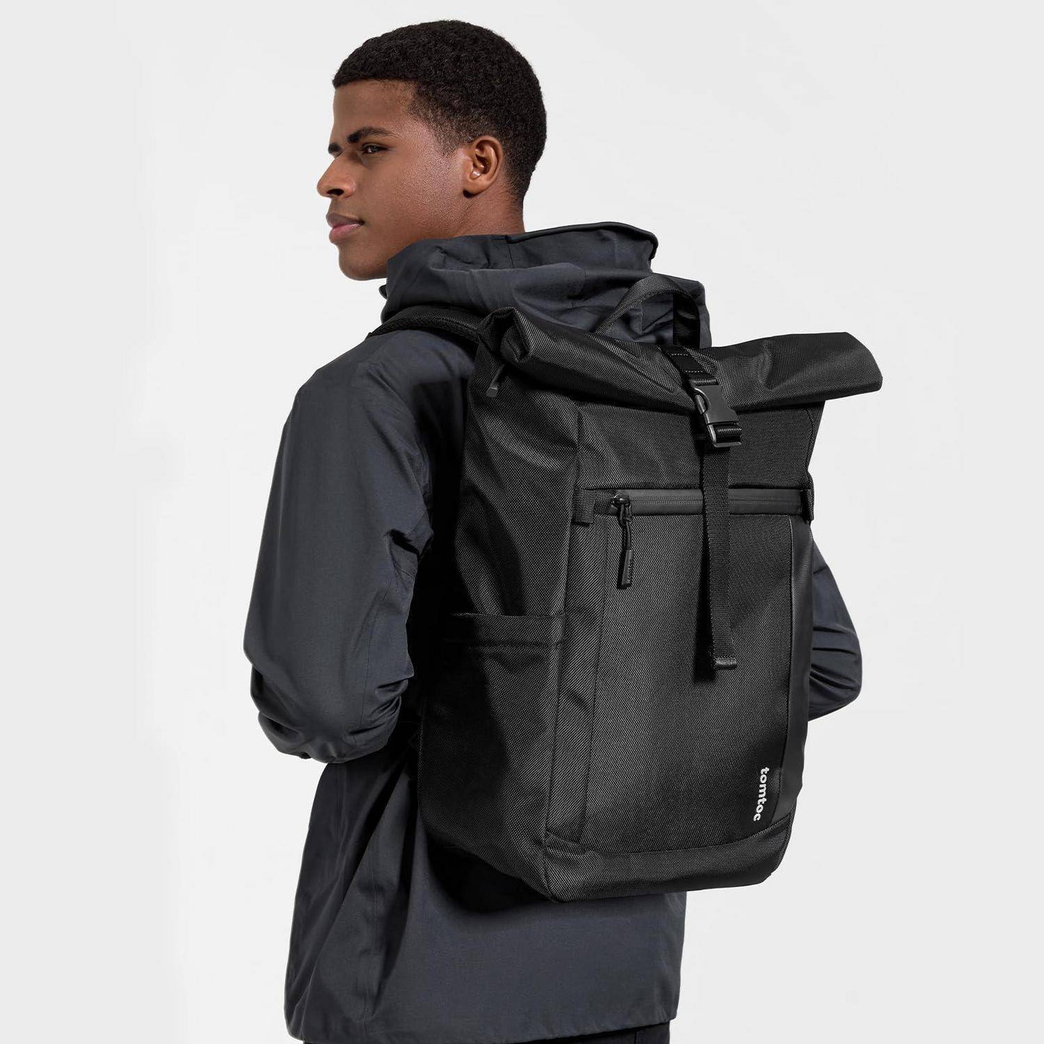 Mann, der einen schwarzen Rollrucksack mit Seitentasche und Frontverschluss trägt, in der Profilansicht vor einem einfachen, hellen Hintergrund dargestellt.
