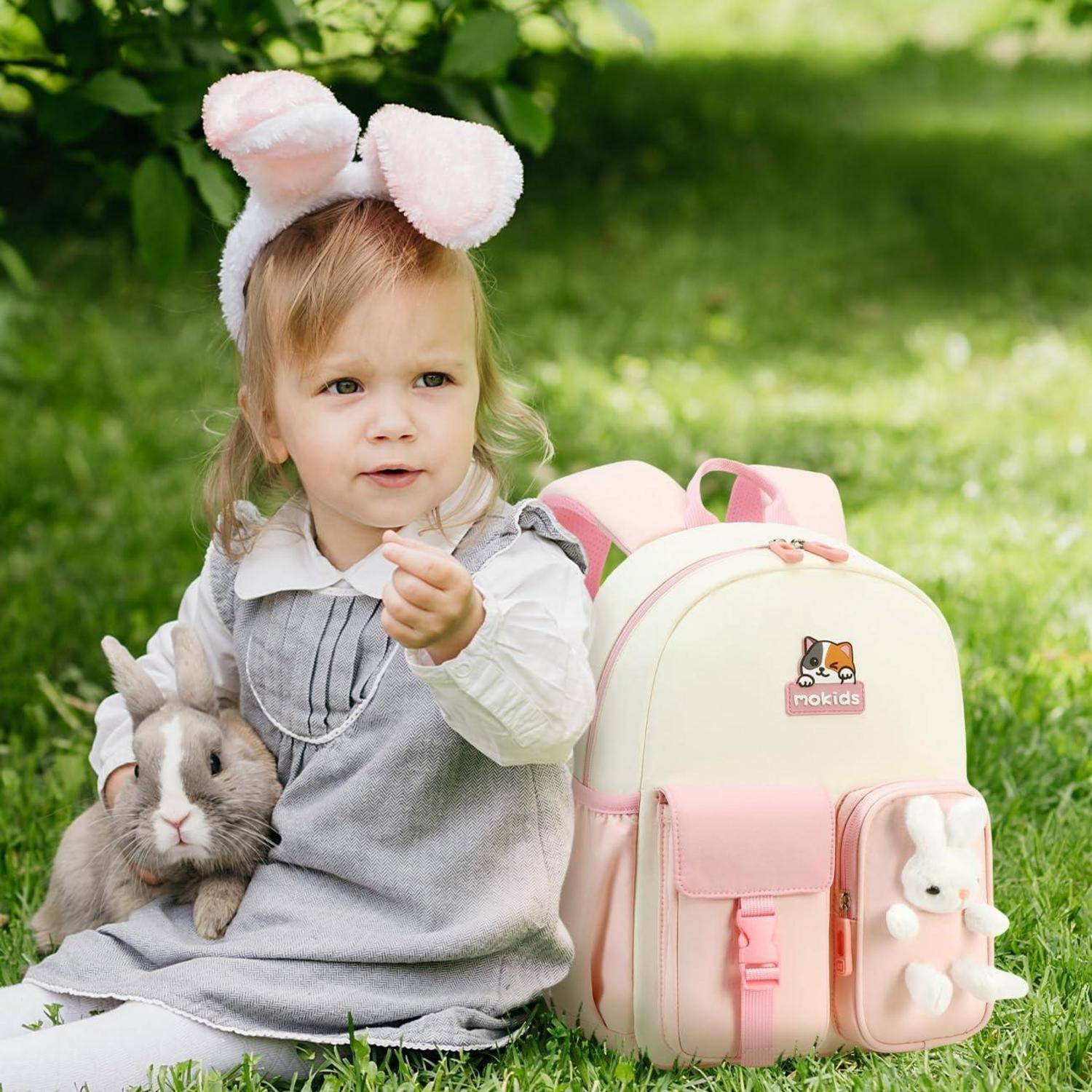 Ein kleines Kind mit Hasenohren sitzt auf dem Gras und hält ein Plüschkaninchen neben einem rosa-cremefarbenen Rucksack.