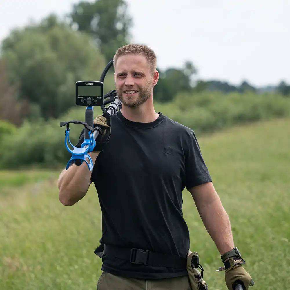 Ein lächelnder Mann im Freien trägt einen Metalldetektor auf der Schulter in einem grasbewachsenen Feld, trägt Handschuhe und ein schwarzes T-Shirt.