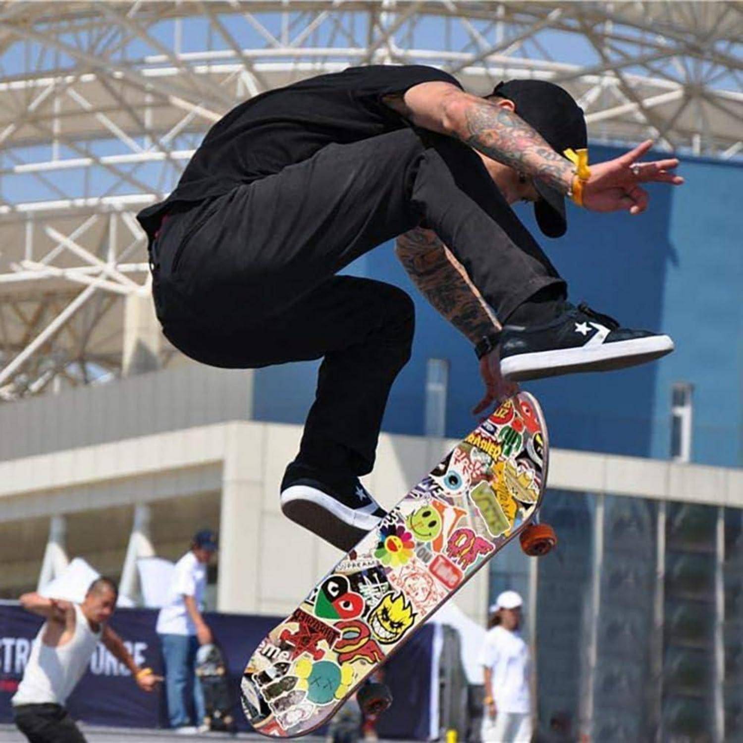 Skateboarder in der Luft, der einen Trick über einem mit Aufklebern bedeckten Skateboard in einem Outdoor-Skatepark ausführt.