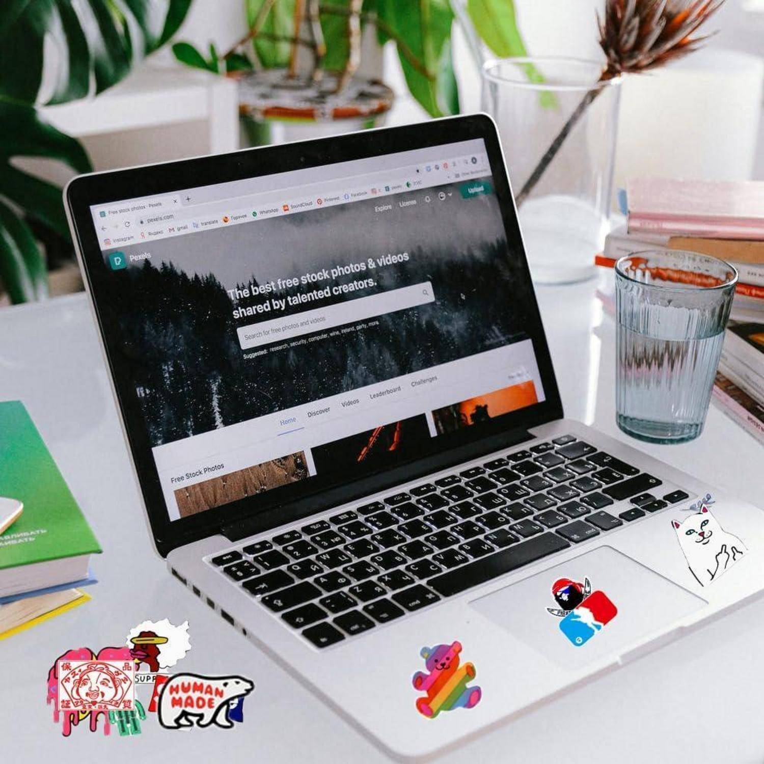 Laptop auf einem Schreibtisch, der eine Homepage einer Stock-Foto-Website zeigt, mit Büchern, einem Glas Wasser und bunten Aufklebern herum.