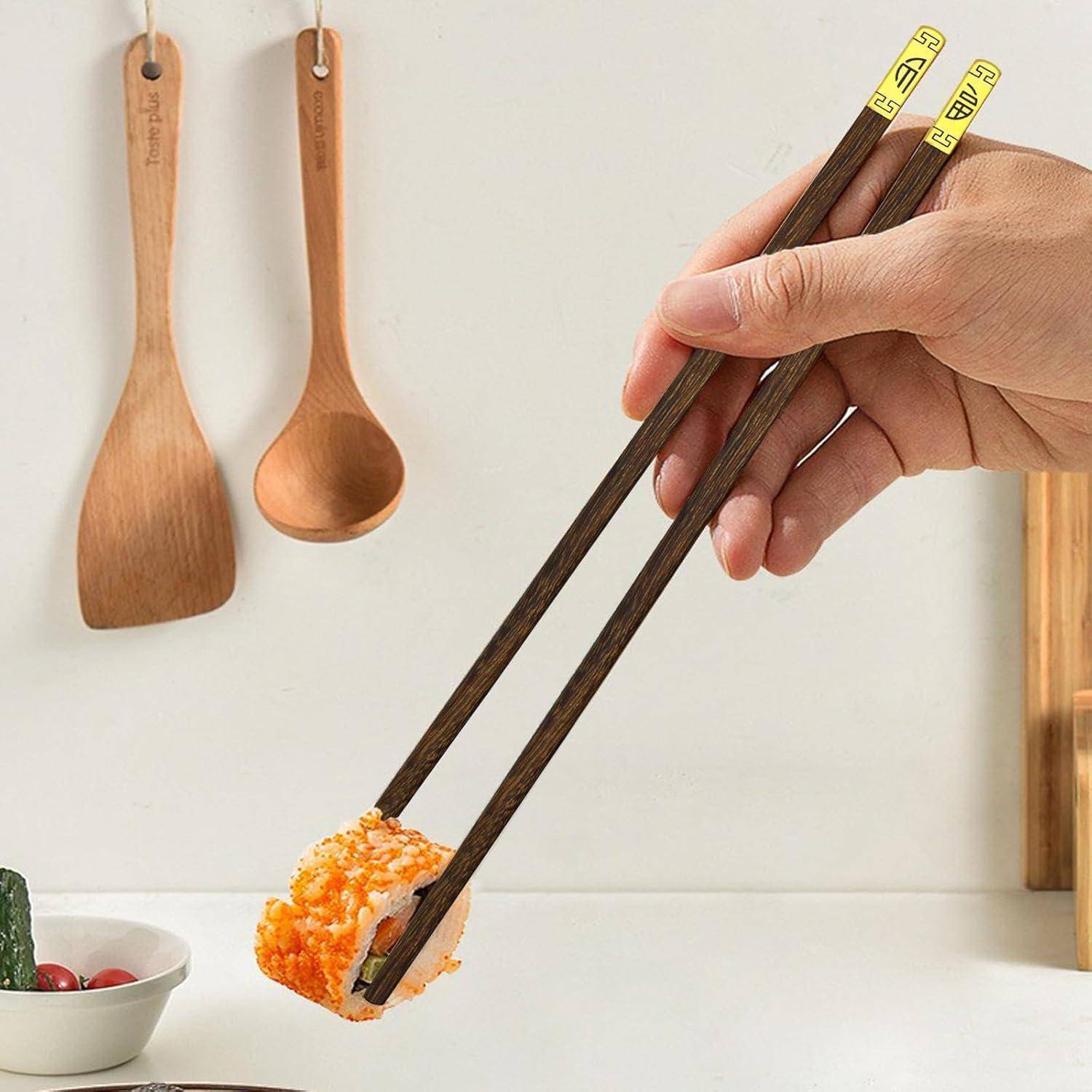 Hand hält dunkle Holzstäbchen, die eine Sushi-Rolle über einer weißen Arbeitsplatte greifen, mit hölzernen Utensilien, die an der Wand im Hintergrund hängen.