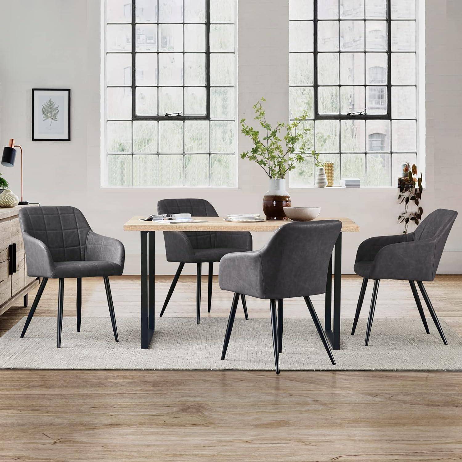Modernes Esszimmer mit einem Tisch aus hellem Holz, vier grau gepolsterten Stühlen, großen Sprossenfenstern und einer Vase mit Grünpflanzen auf dem Tisch.