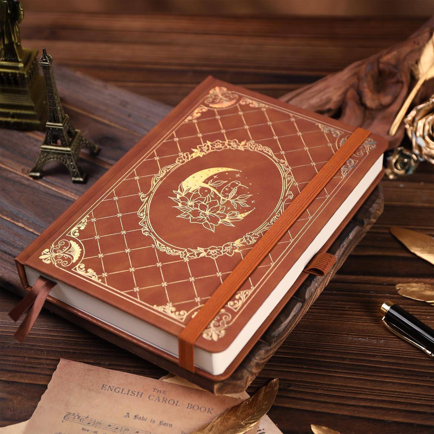 Ein dekoratives braunes Hardcover-Journal mit goldenem Mond- und Blütenprägung und elastischem Band, das auf einem Holzschreibtisch mit Vintage-Dekoration ruht.