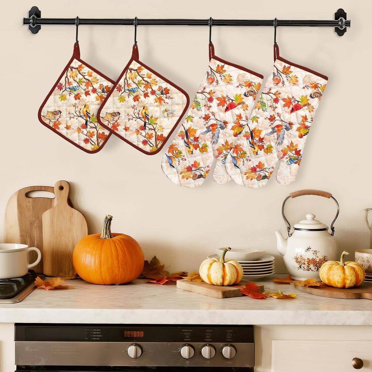 Herbstliche Küchendisplay mit Ofenhandschuhen im Herbstblatt-Muster, Topflappen, Kürbissen und einem blumigen Wasserkocher auf einer Arbeitsplatte über einem Herd.