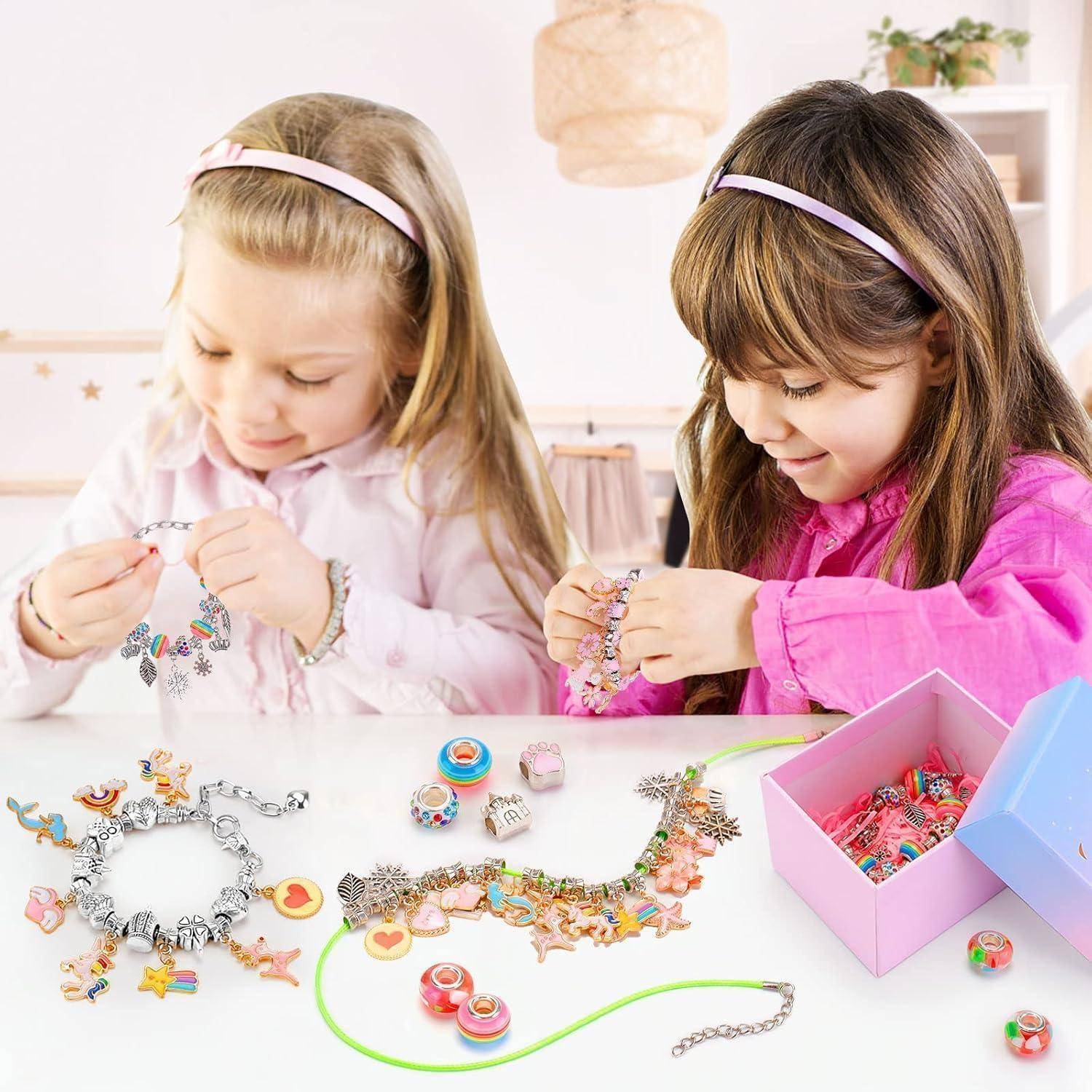 Zwei junge Mädchen stellen bunte Charm-Armbänder an einem Tisch her, mit Perlen, fertigem Schmuck und einer offenen Schachtel mit Bastelmaterialien.