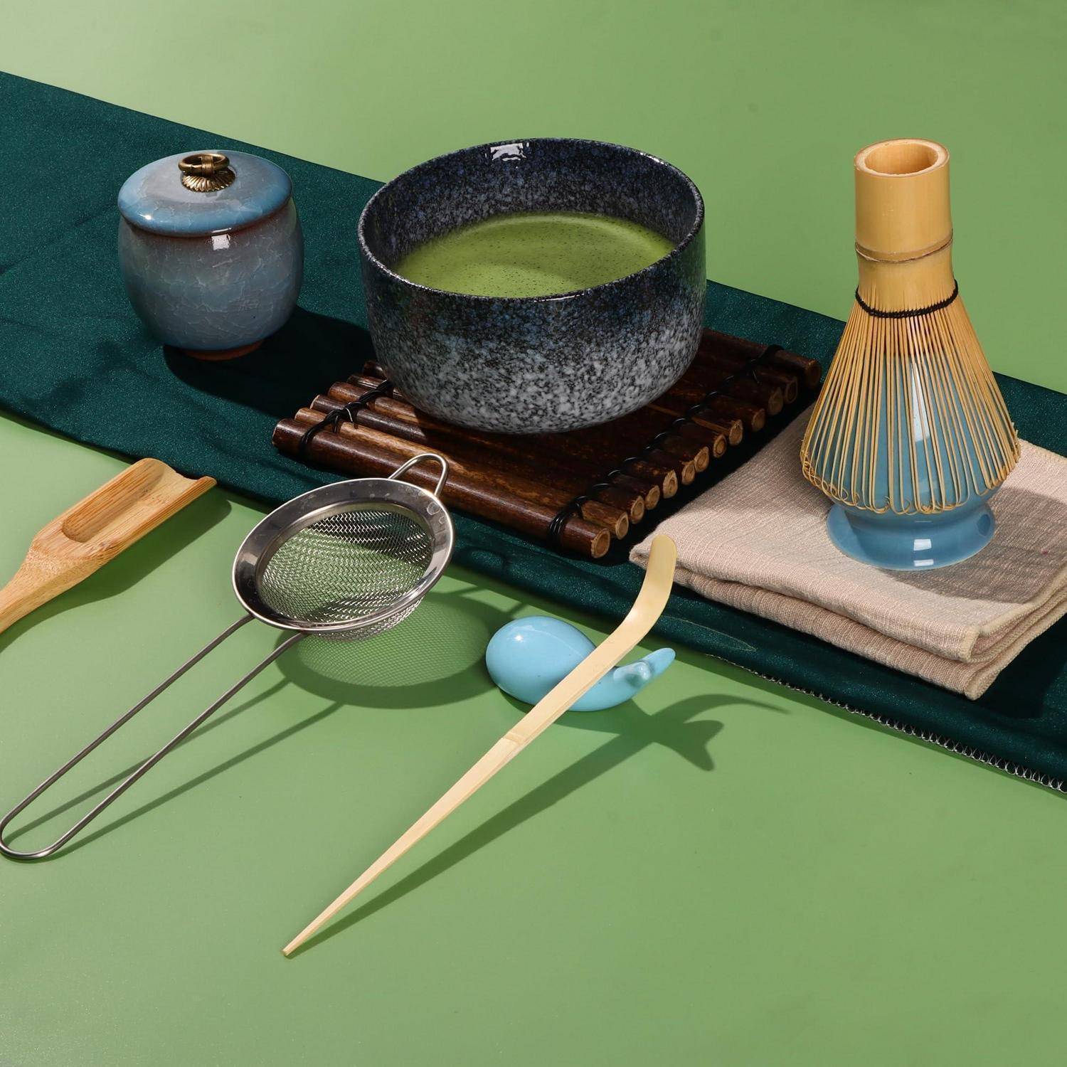 Matcha-Teeset mit einer Schale grünem Tee, Schneebesen, Sieb, Löffel und Keramikbehältern auf einer grünen Oberfläche.