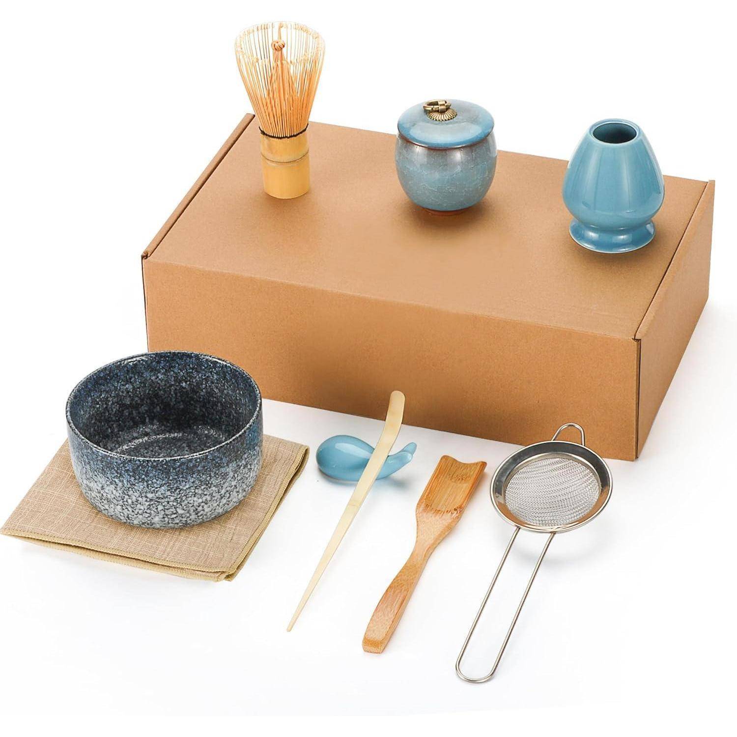 Matcha-Teezeremonie-Set mit Schale, Schneebesen, Löffel, Sieb, Tasse und Keramikgefäßen, angeordnet auf und um eine Kartonschachtel.