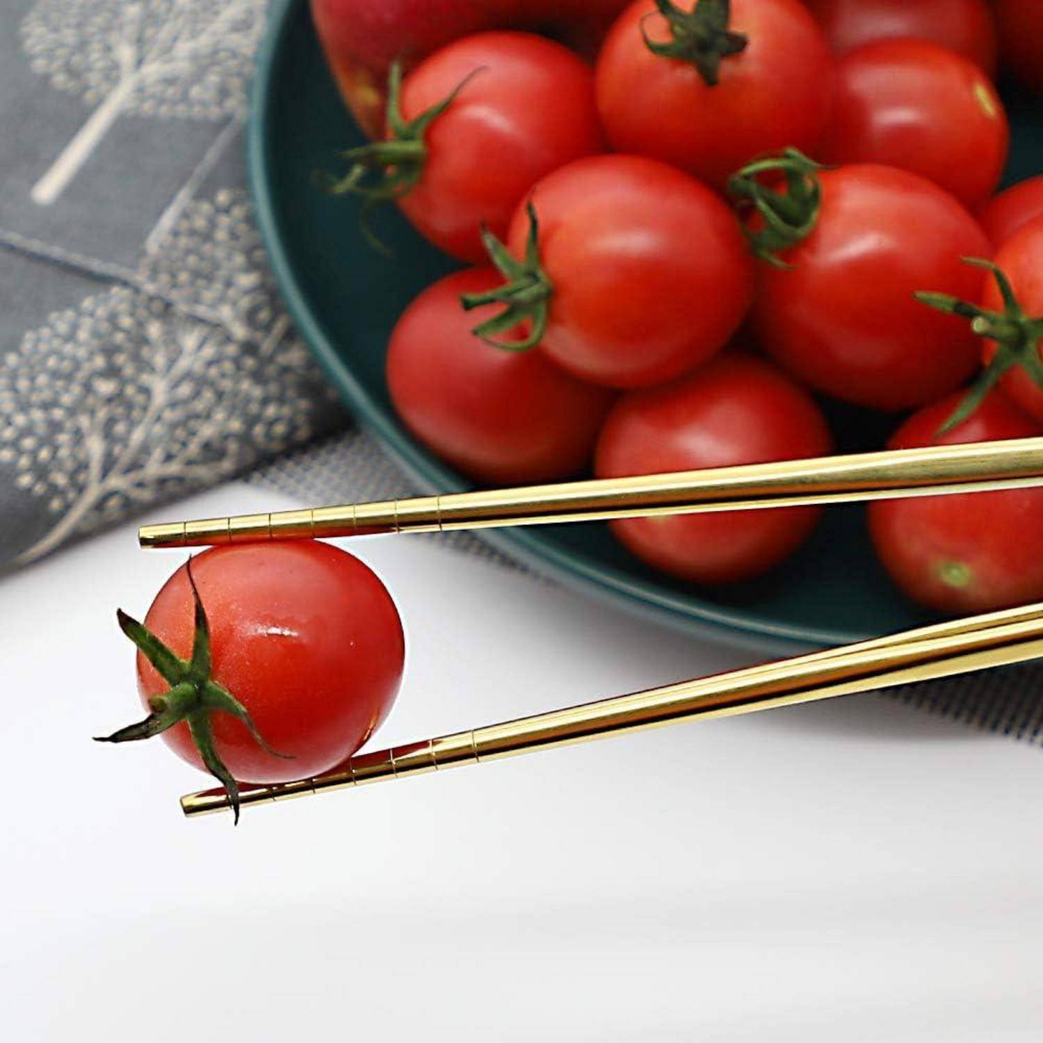 Golde Essstäbchen halten eine Kirschtomate neben einer Schüssel mit Tomaten auf einer weißen Oberfläche.