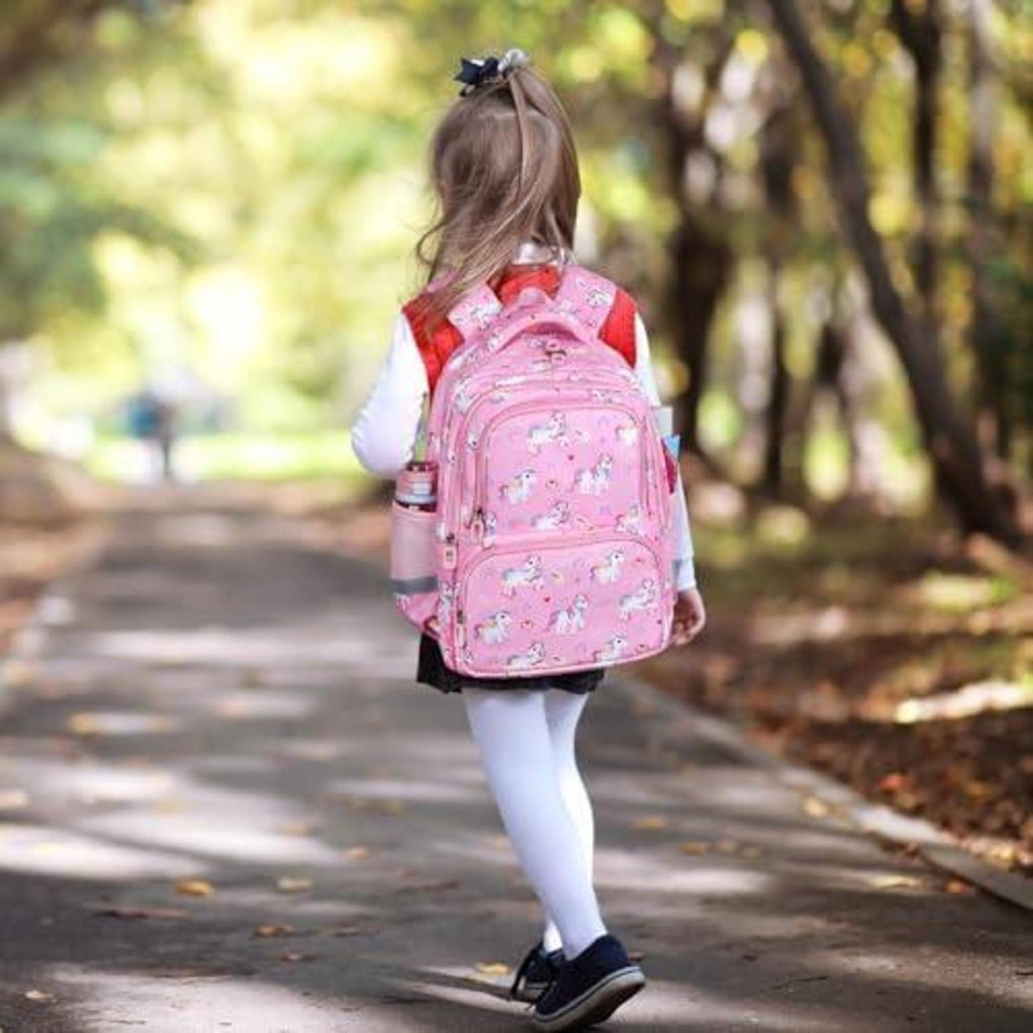 Junges Mädchen, das auf einem baumgesäumten Pfad weggeht, trägt einen rosa Rucksack und weiße Strumpfhosen, während Sonnenlicht durch die Bäume filtert.