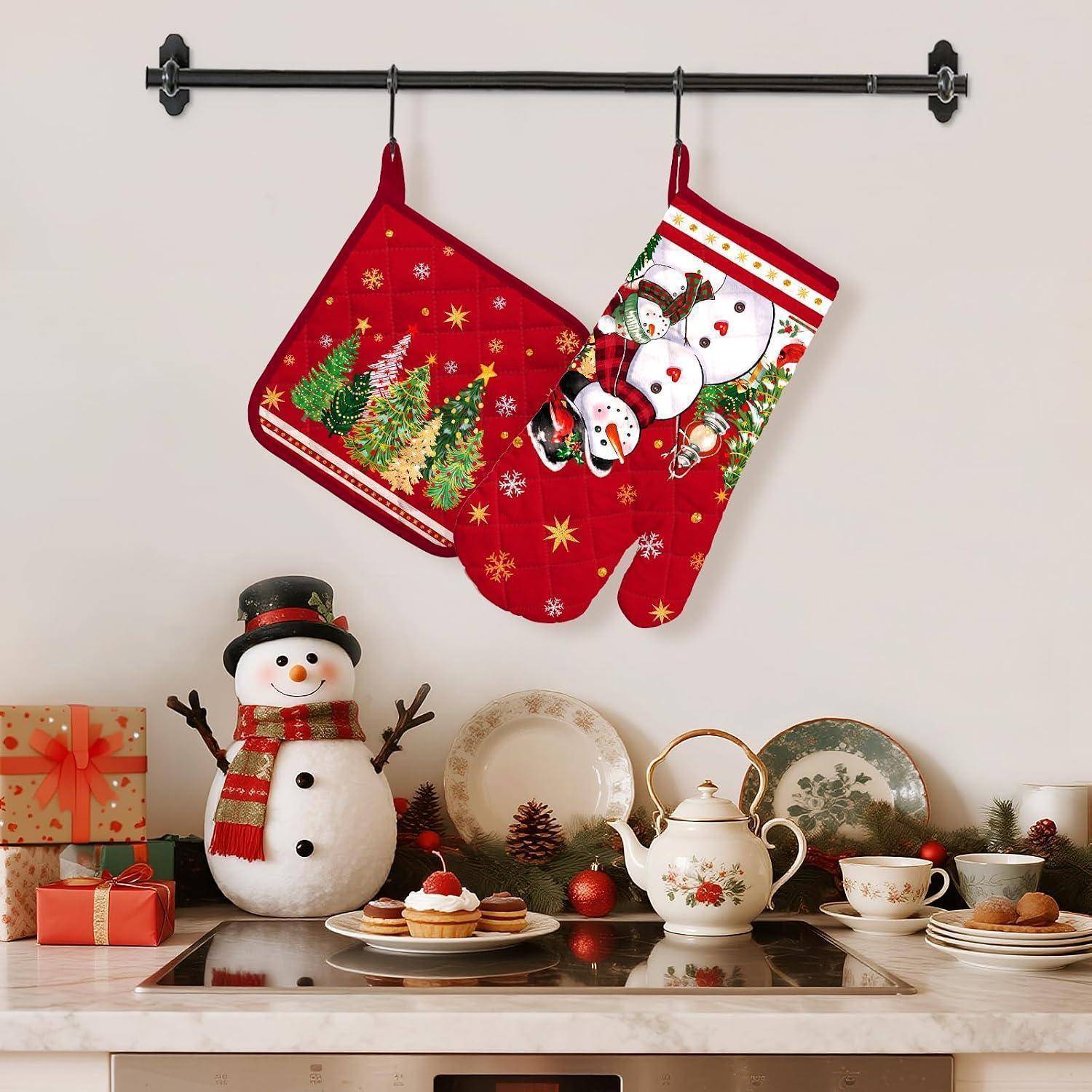 Zwei rote Weihnachtsküchenhandschuhe hängen über einer festlichen Küchendisplay mit einer Schneemann-Dekoration, einer Teekanne, Teetassen, Tellern, Geschenken und Christbaumkugeln.