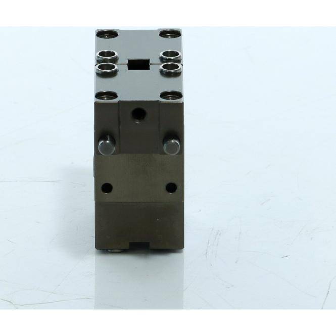 Ein kleines schwarzes rechteckiges Metallteil mit mechanischer Funktion, mehreren Schraubenlöchern und einem zentralen Schlitz, aufrecht auf einer hellen Oberfläche stehend.