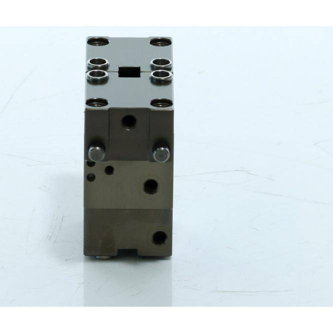 Kleiner metallischer mechanischer Block mit mehreren gebohrten Löchern und zwei seitlichen Stiften, aufrecht auf einer hellen Oberfläche stehend.