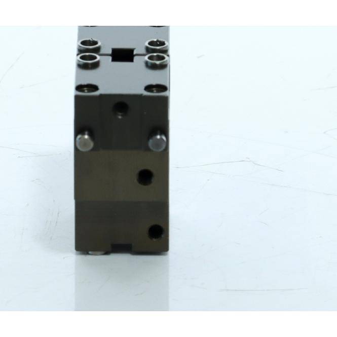 Ein schwarzer Metallblock aus Kunststoff mit rechteckiger Form, mehreren Schraubenlöchern und Stiften, aufrecht auf einer weißen Oberfläche stehend.