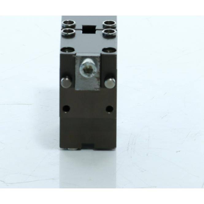 Schwarzer Metallmechanischer Block mit vier Schraubenlöchern oben und einer zentralen Schraube, aufrecht vor weißem Hintergrund dargestellt.