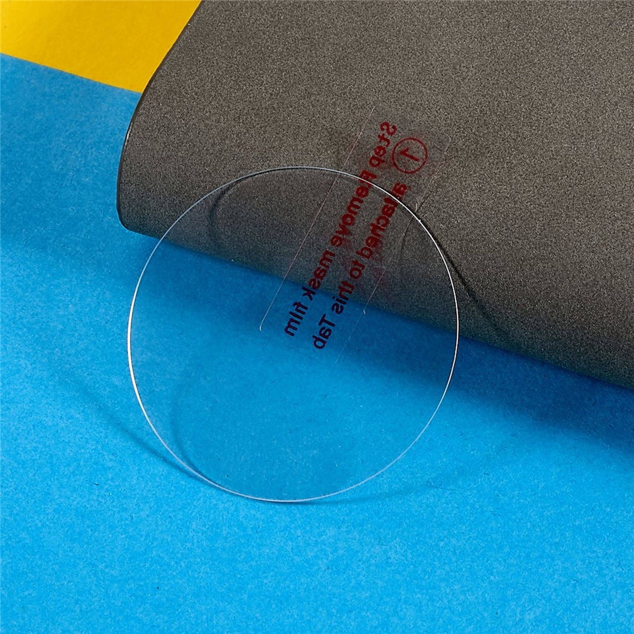 Klare runde Kunststofflinse oder Scheibe auf blauem und gelbem Papier neben einem grau gerollten Blatt mit rot gedrucktem Text.
