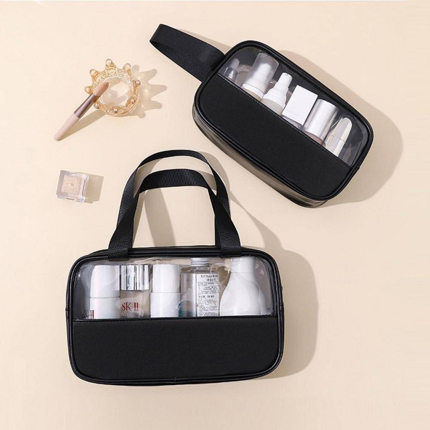 Zwei schwarze transparente Kulturtaschen mit Griffen, jede mit Travel-Size-Flaschen und Tuben gefüllt, auf einer beigen Oberfläche mit einer Haarspange und einem kleinen Behälter in der Nähe.