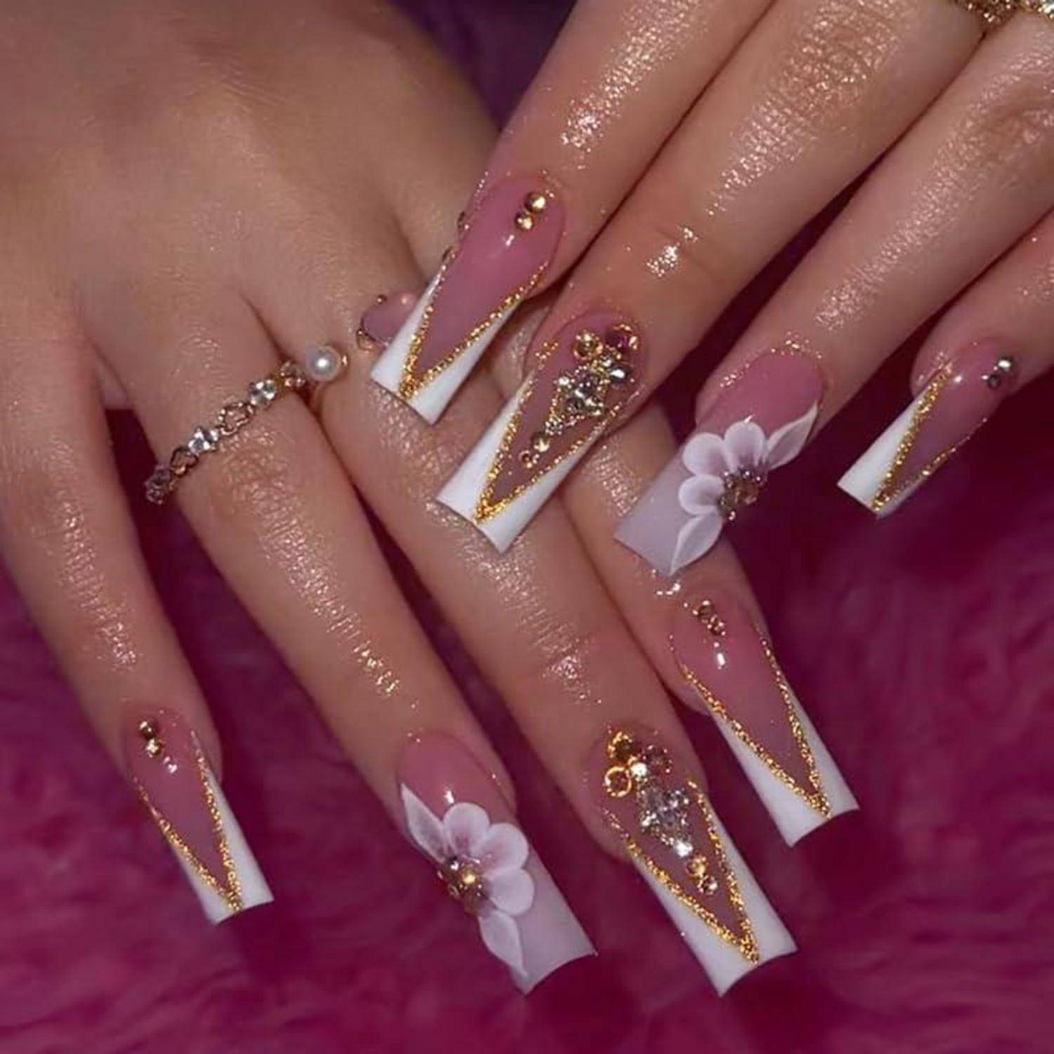 Nahaufnahme von Händen mit langen rosa-weißen Acrylnägeln mit goldenen Glitzereffekten, Strasssteinen und kleinen weißen Blütendesigns.