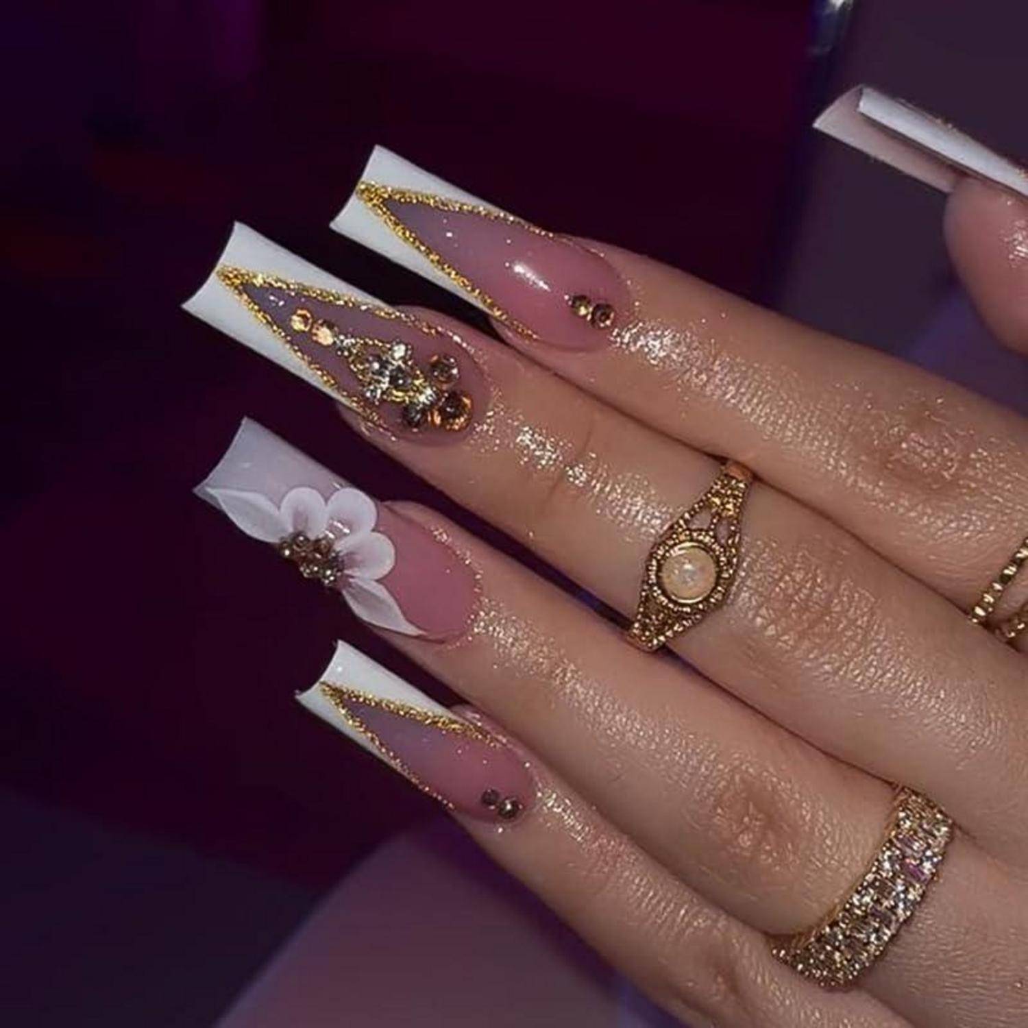 Nahaufnahme einer manikürierten Hand mit langen rosa-weißen Sargnägeln, goldenen Glitzereffekten, Blumennailart und mehreren goldenen Ringen.