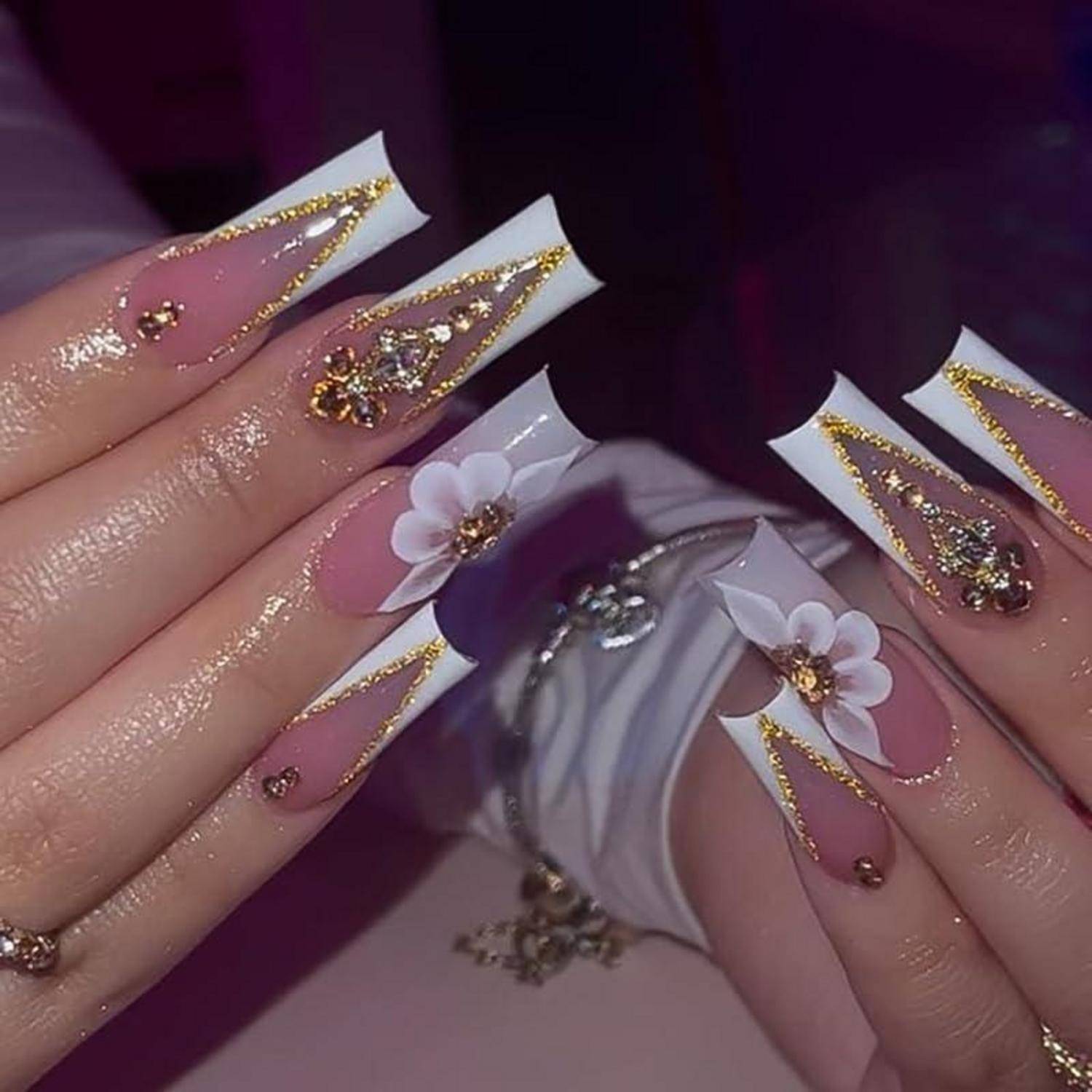 Nahaufnahme von langen, rosa-weiß gefärbten Acrylnägeln mit Goldglitter, Juwelen-Akzenten und 3D-weißen Blumenverzierungen auf beiden Händen.