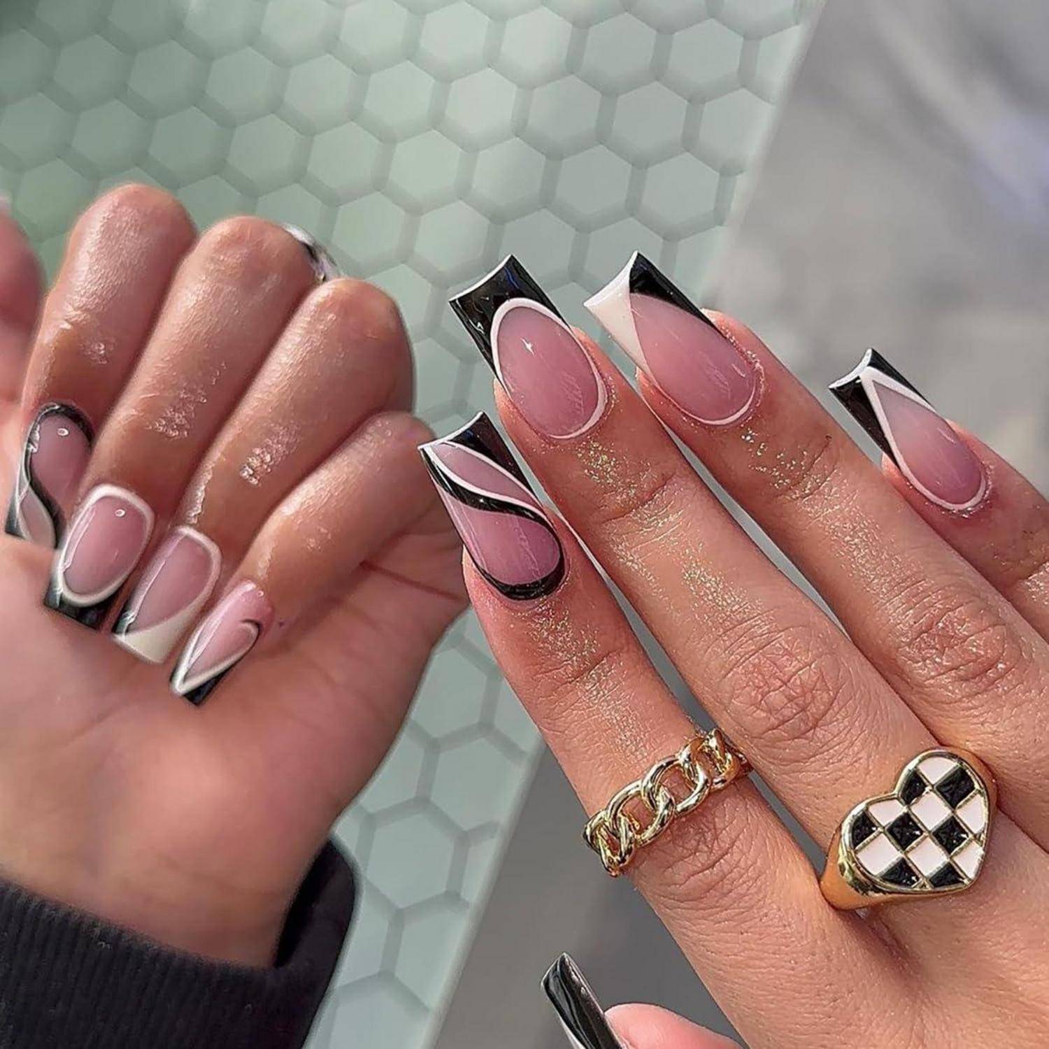 Nahaufnahme von Händen mit langen Acrylnägeln in rosa Ombré mit schwarzweißen abstrakten Spitzen sowie Goldringen.