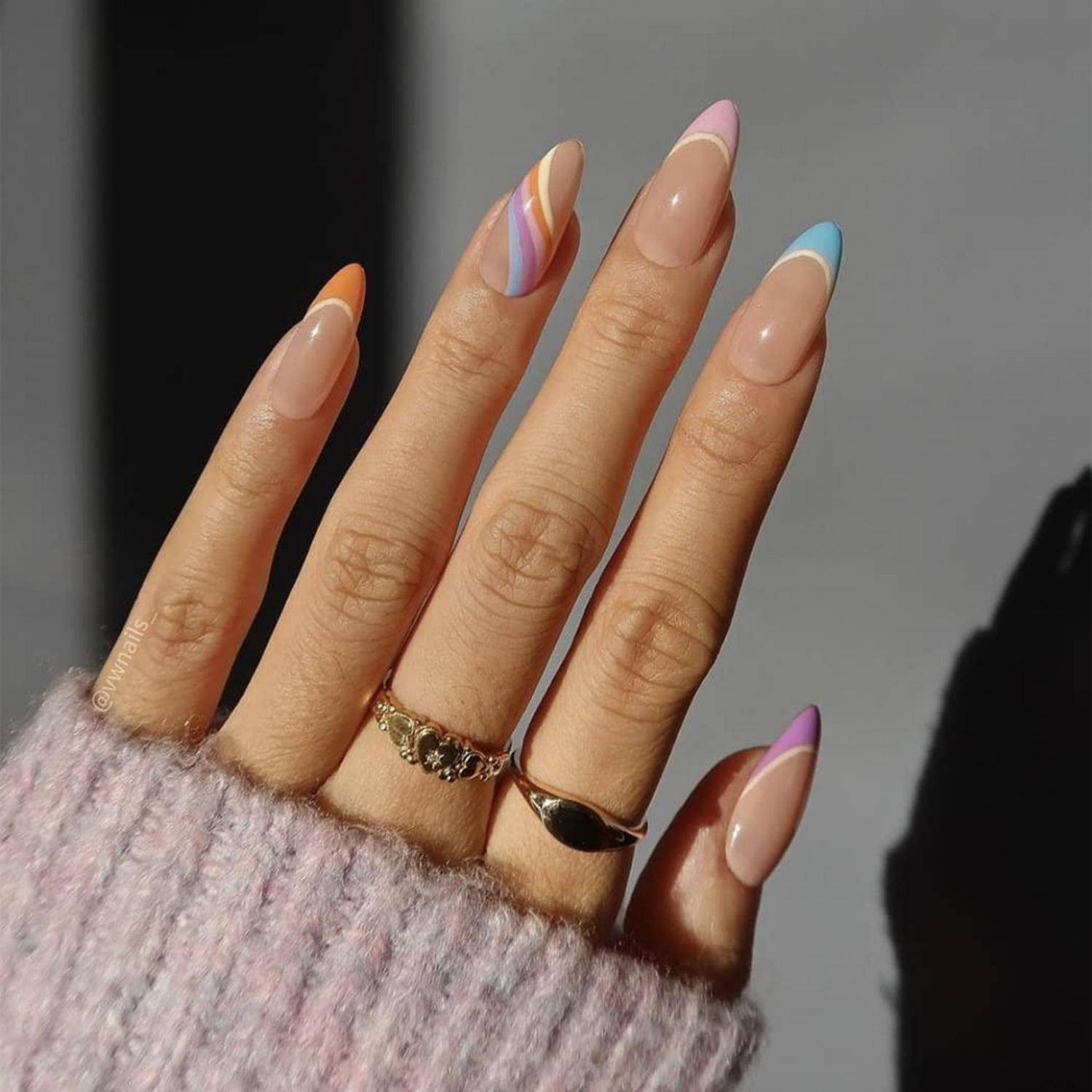 Hand mit langer pastellfarbener französischer Nagelspitzenmanikure und goldenen Ringen, teilweise von einem flauschigen rosa Ärmel bedeckt.