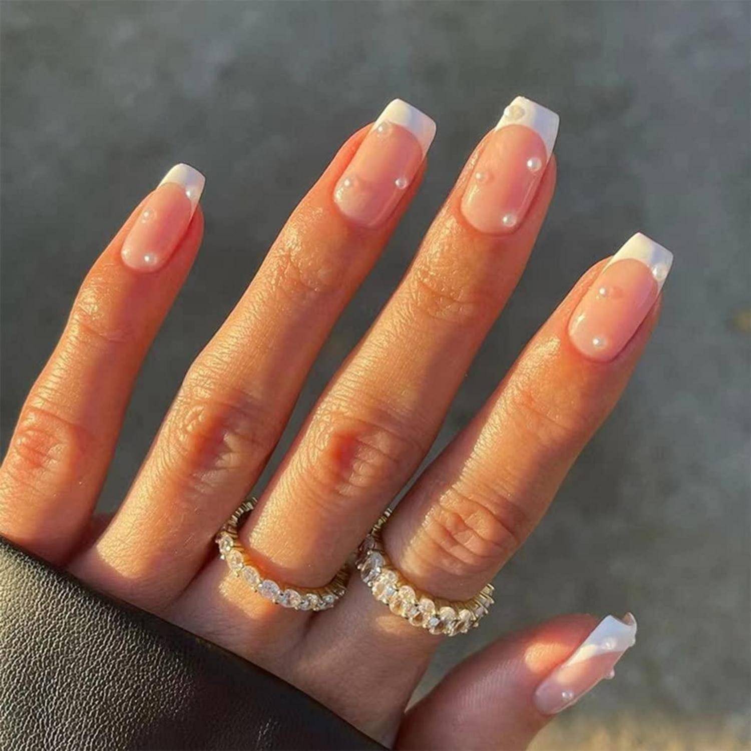Hand mit langer French-Ombré-Maniküre und Perlakzenten, die zwei goldene Edelsteinringe trägt.